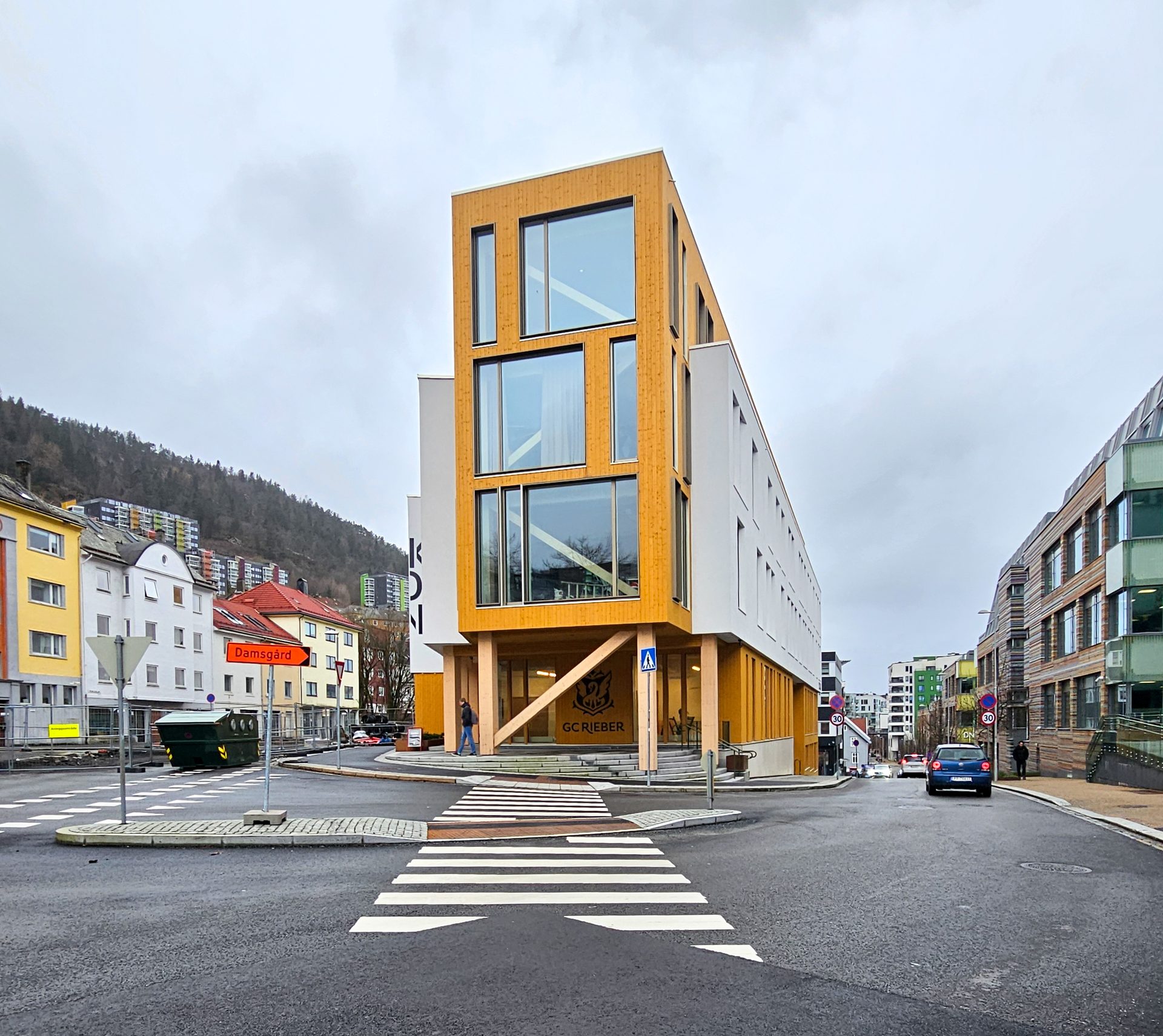 Modern wooden building on a street corner with crosswalk, other buildings, and a wooded hill.