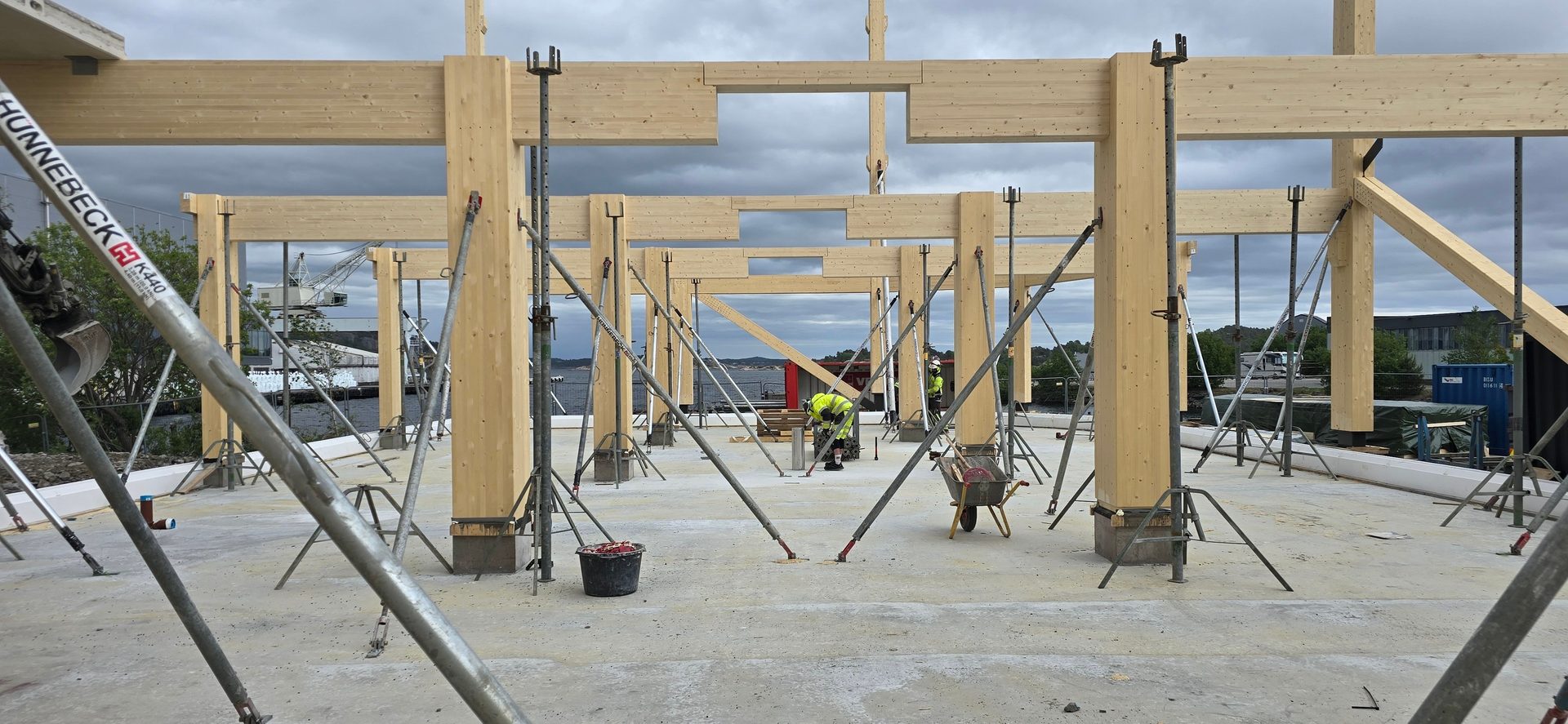 Timber frame building under construction with metal props, and a port in the background.