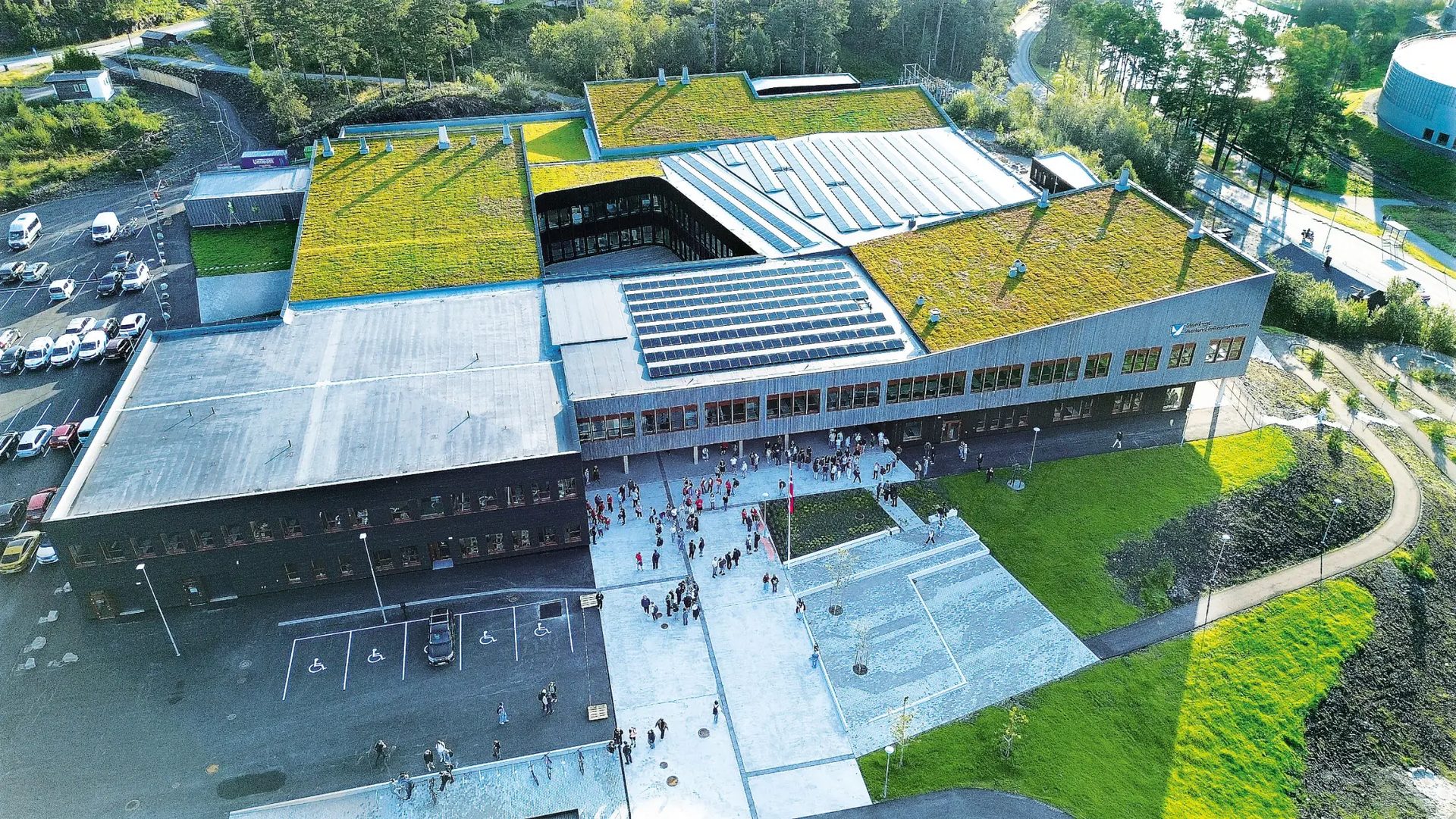 Aerial view of a modern school building with extensive green roofs, solar panels, and a plaza bustling with students.