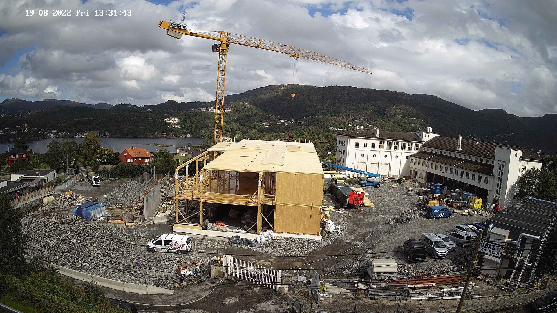 Construction site with a wooden building, crane, white buildings, a lake, and mountains under cloudy skies.