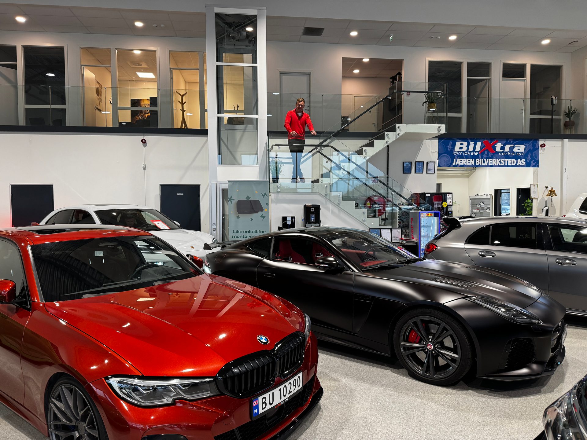 New red BMW and black Jaguar cars in a spacious dealership showroom with a man on the stairs.