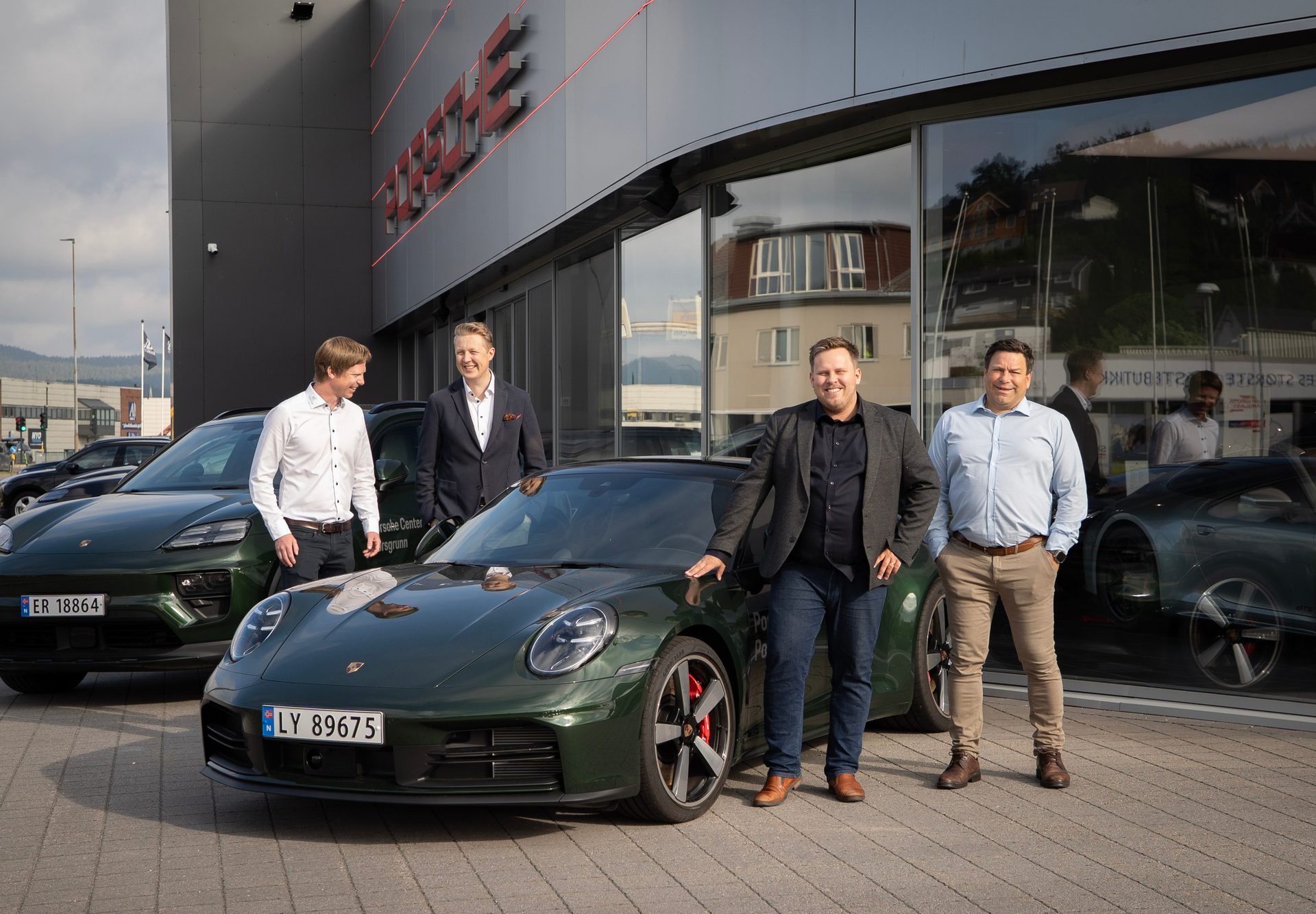 Four men pose with two green Porsche cars outside a dealership.