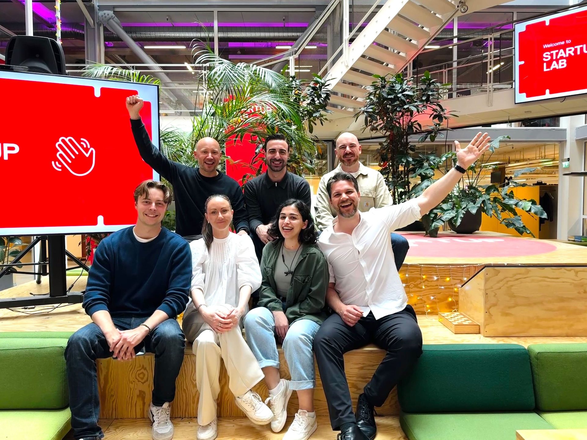 Seven smiling people pose in a modern space with "STARTUP LAB" on a screen behind them.