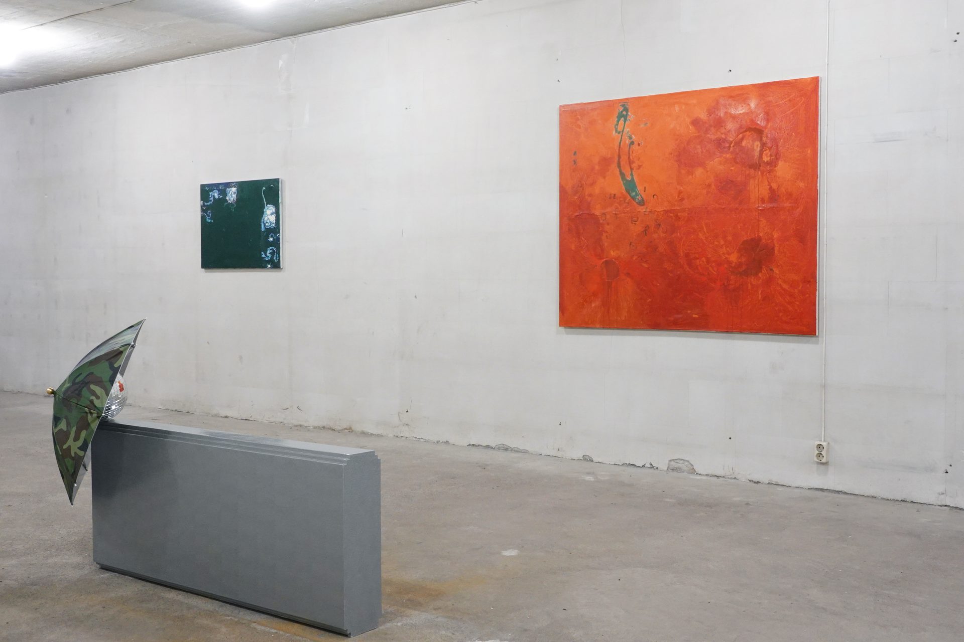Two abstract paintings (green, orange) and an umbrella on a plinth in a gallery space.