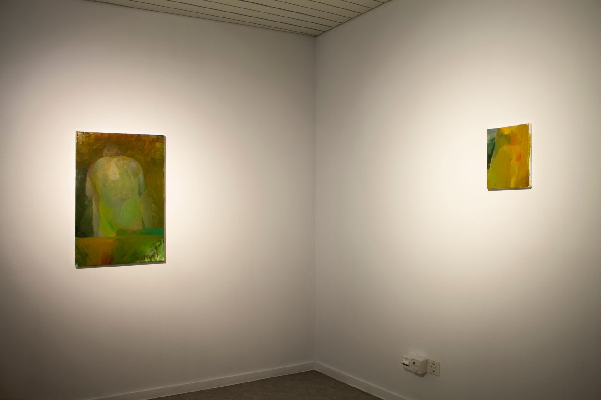Two abstract green/yellow paintings of different sizes on white gallery walls in a corner.