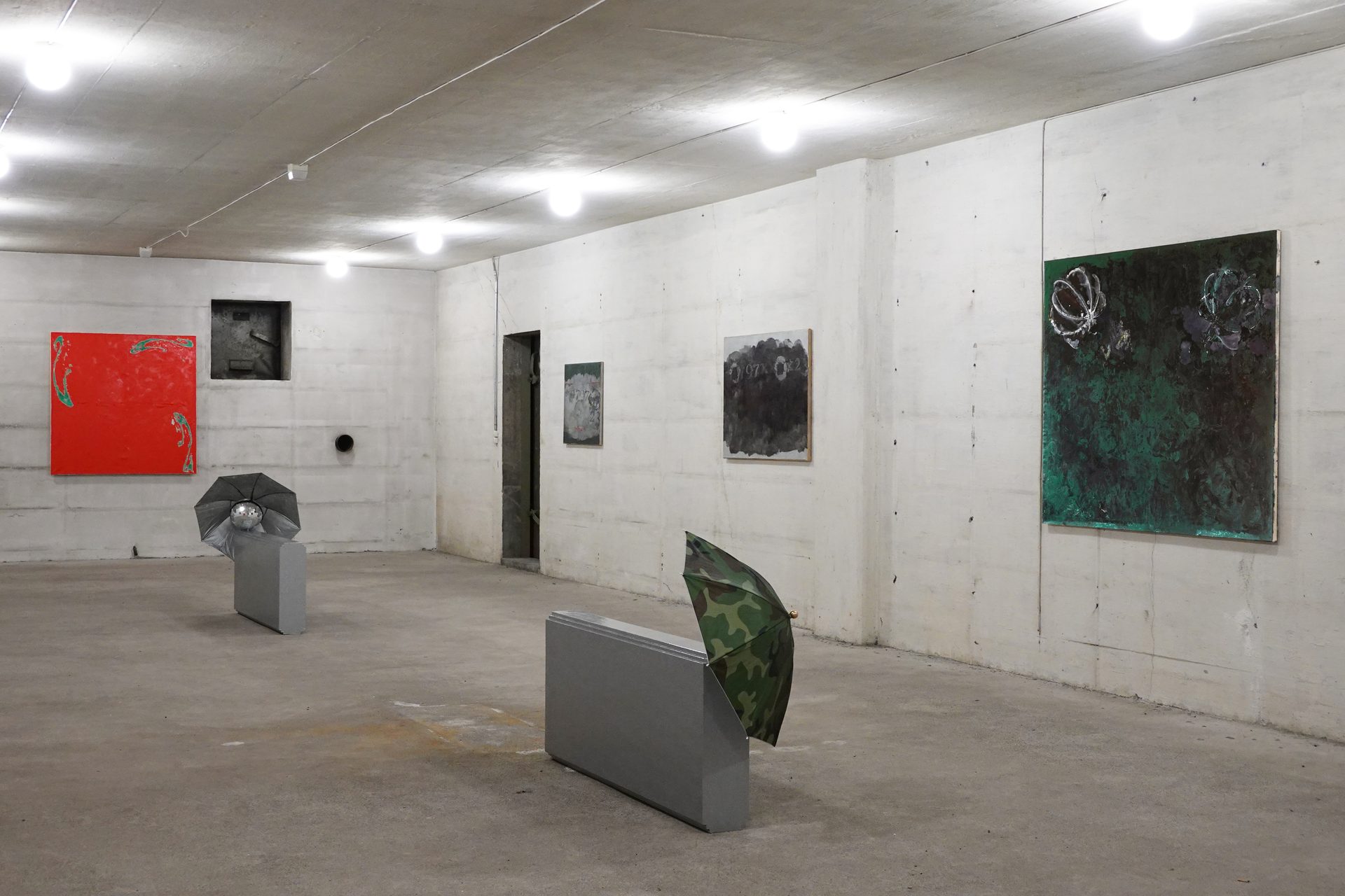 An art gallery with abstract paintings on concrete walls, featuring two unique umbrella sculptures.