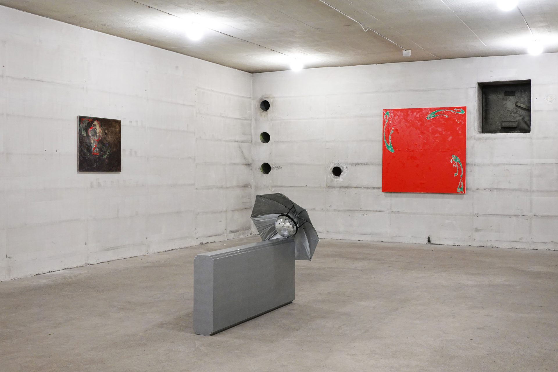 An art installation in a raw concrete room featuring two paintings, a sculpture with a disco ball, and wall cutouts.