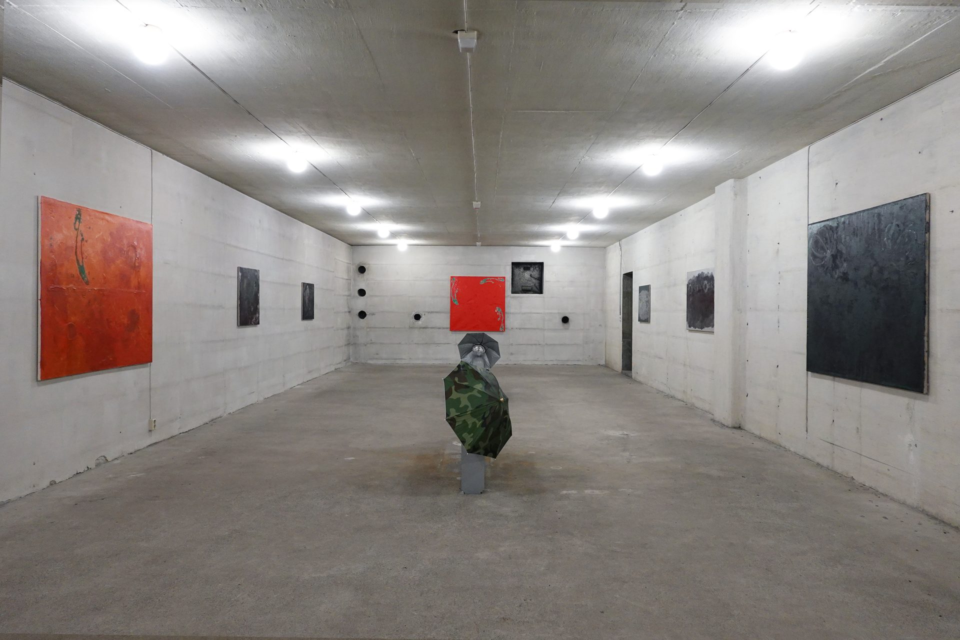 A concrete gallery hall with various paintings on the walls and a camouflage umbrella sculpture.