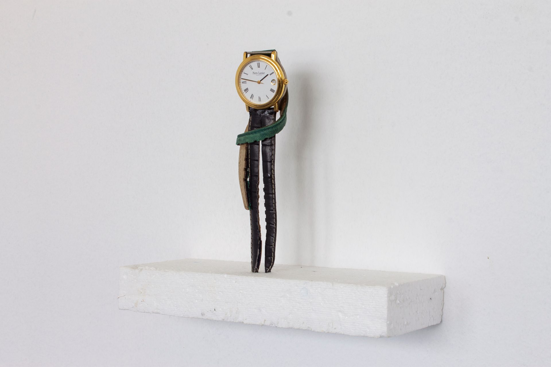 A gold watch with a white face and dark and green straps stands upright on a white block.