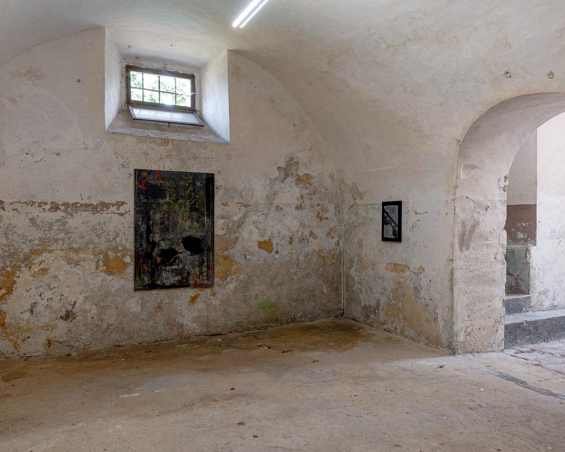 An old, arched room with peeling walls, a window, two abstract paintings, and an arched doorway.