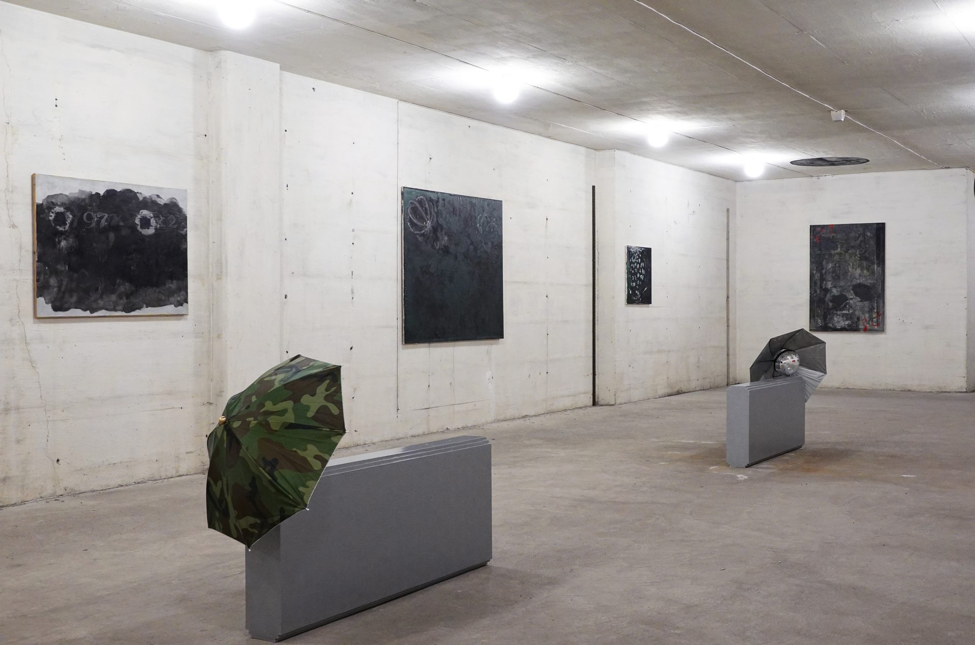 Art gallery with abstract paintings and two umbrella installations on concrete walls and floor.