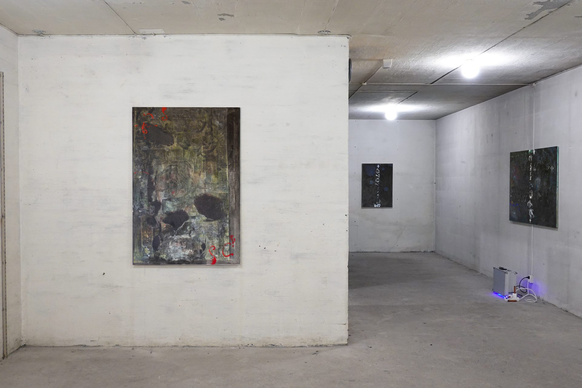 An industrial gallery space showcasing several abstract paintings on concrete walls. A silver box with a blue light is on the floor.