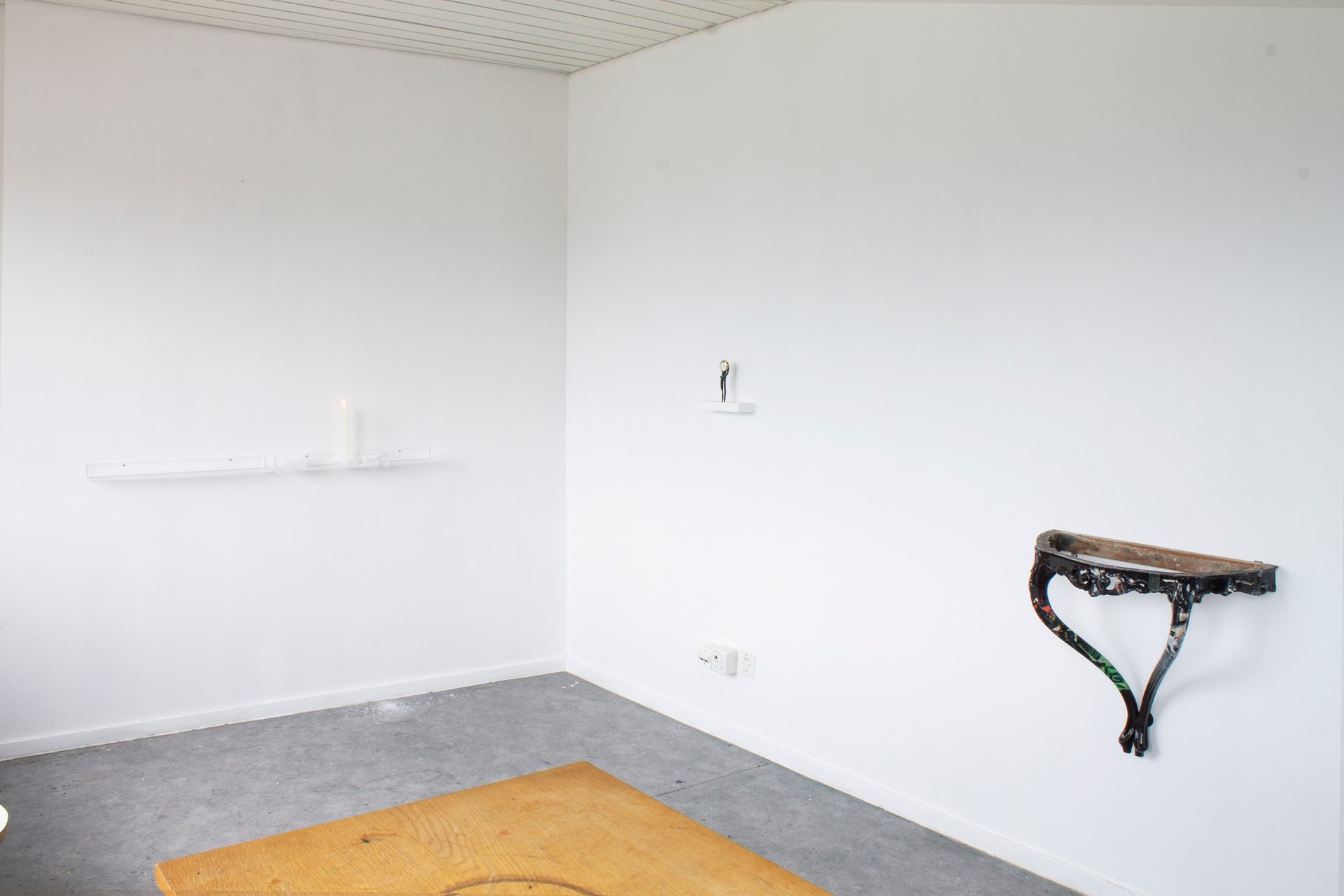 White room with concrete floor, wooden table, burning candle, small figure, and ornate wall shelf.