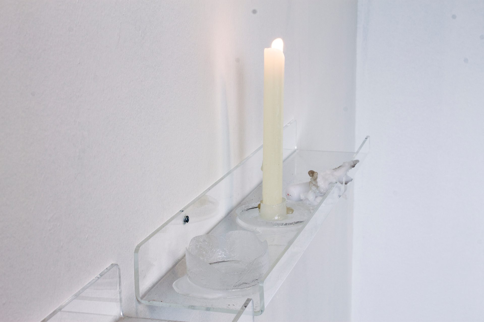A lit candle and clear decorative objects on a transparent shelf mounted on a minimalist white wall.