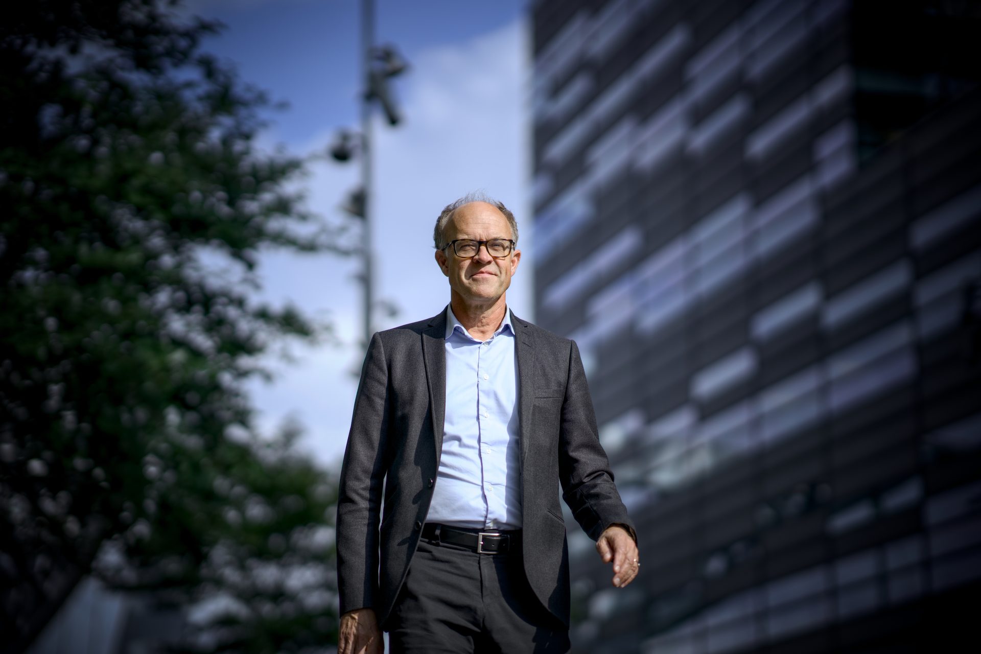 A man in a suit and glasses stands outdoors, with a modern building and trees in the background.