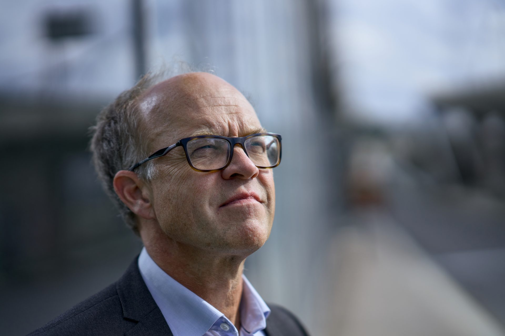 Close-up of a man in glasses and suit jacket looking upwards, with a blurred outdoor background.