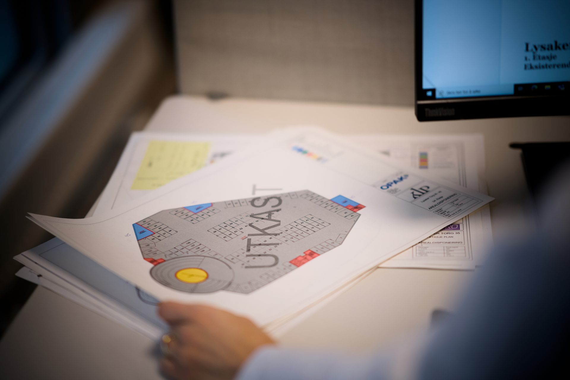 Hand holding a printed floor plan with 'UTKAST' on a desk, a computer screen behind.
