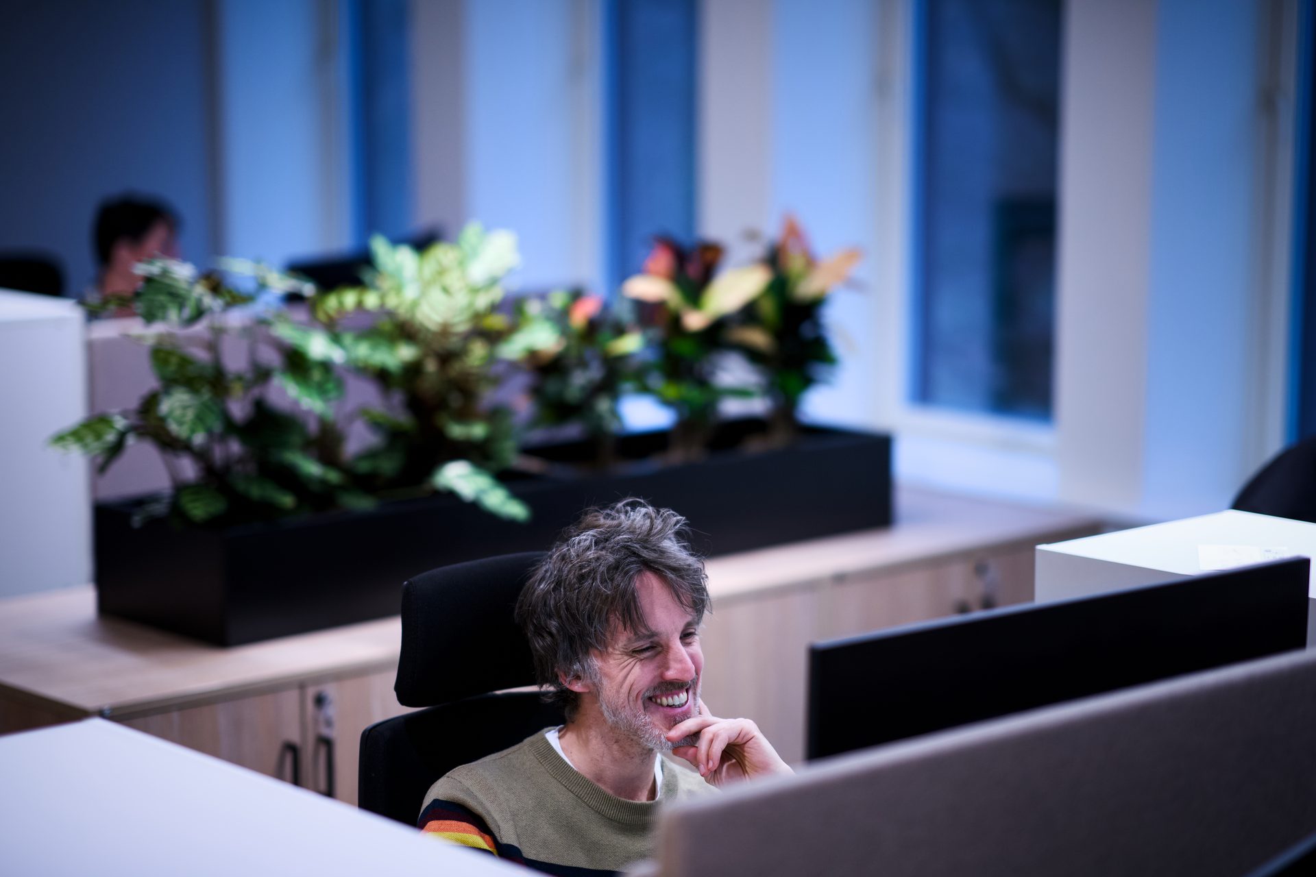 Man laughs at screen in modern office with plants.