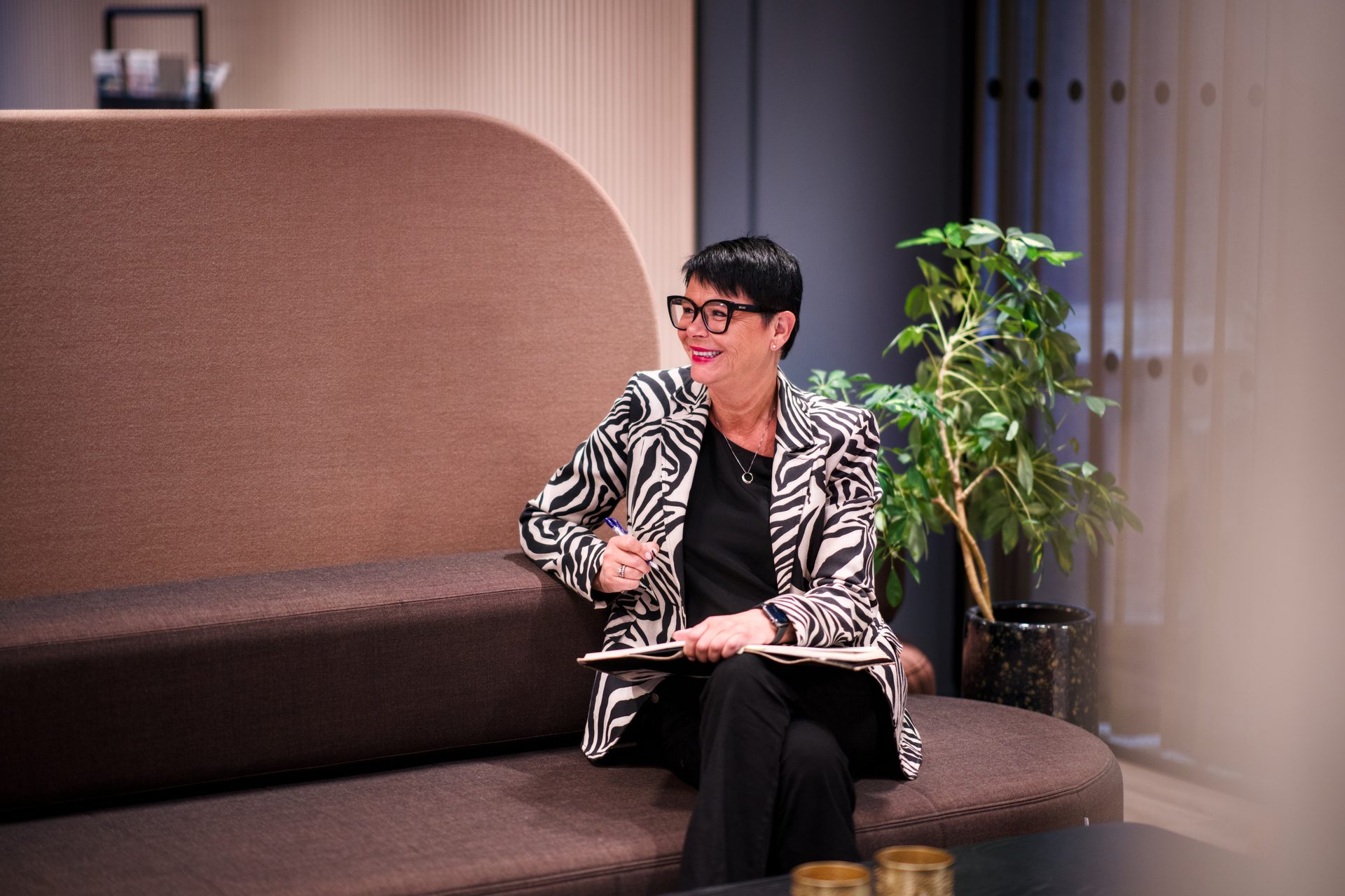 Smiling woman in zebra jacket, glasses, holding a pen and notebook, sitting on a brown sofa.