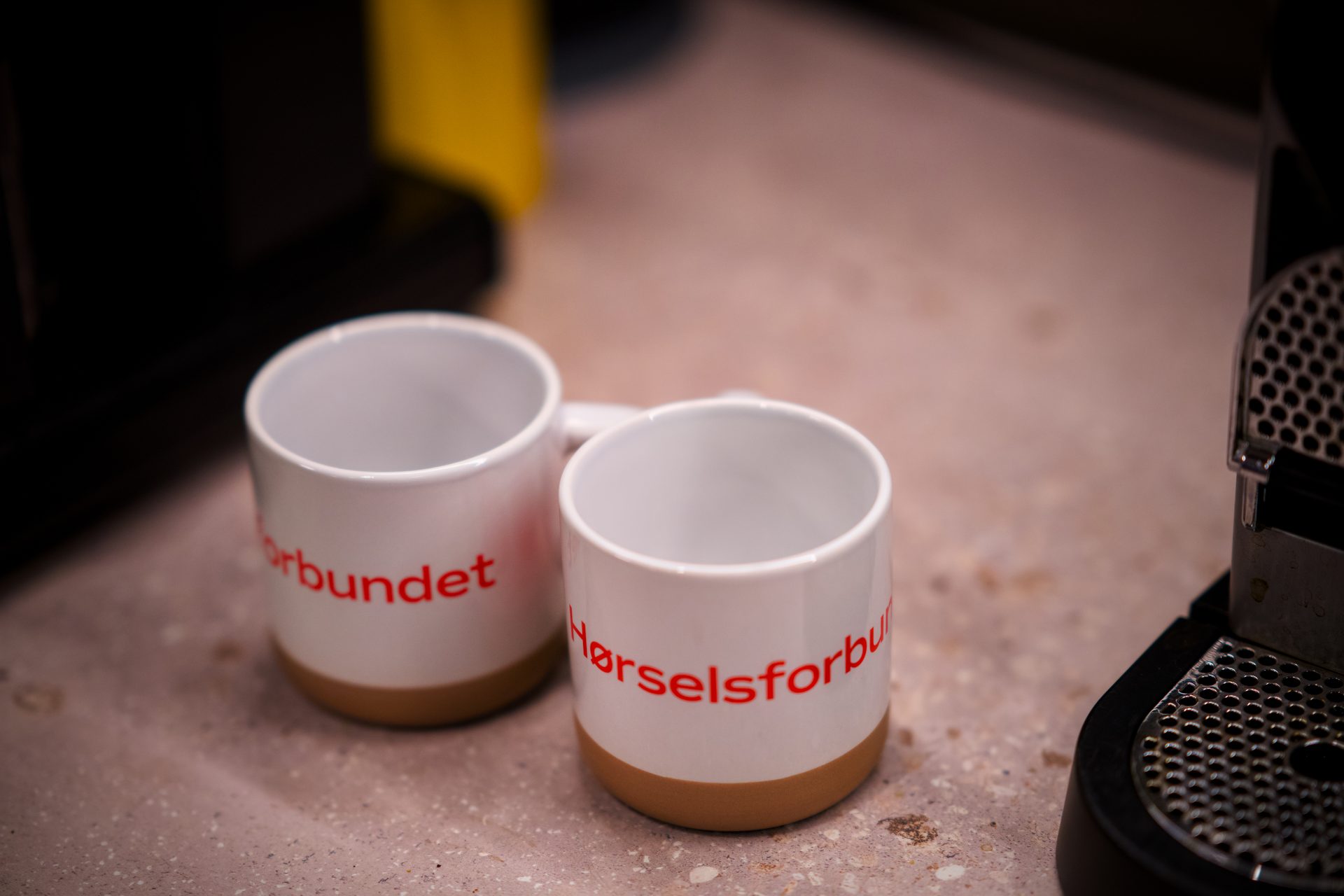 Two white mugs with "Hørselsforbundet" text on a counter next to a coffee machine.