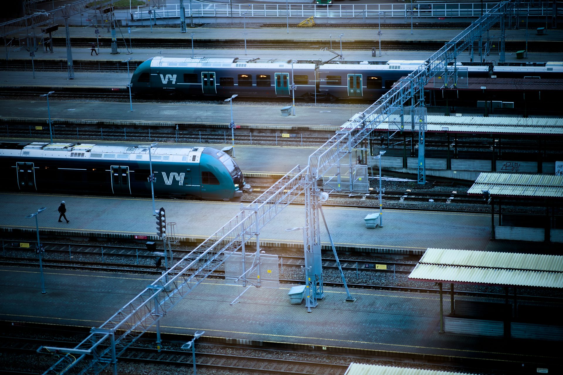 High-angle view of a train station with two green and black Vy trains, tracks, platforms, and people walking.