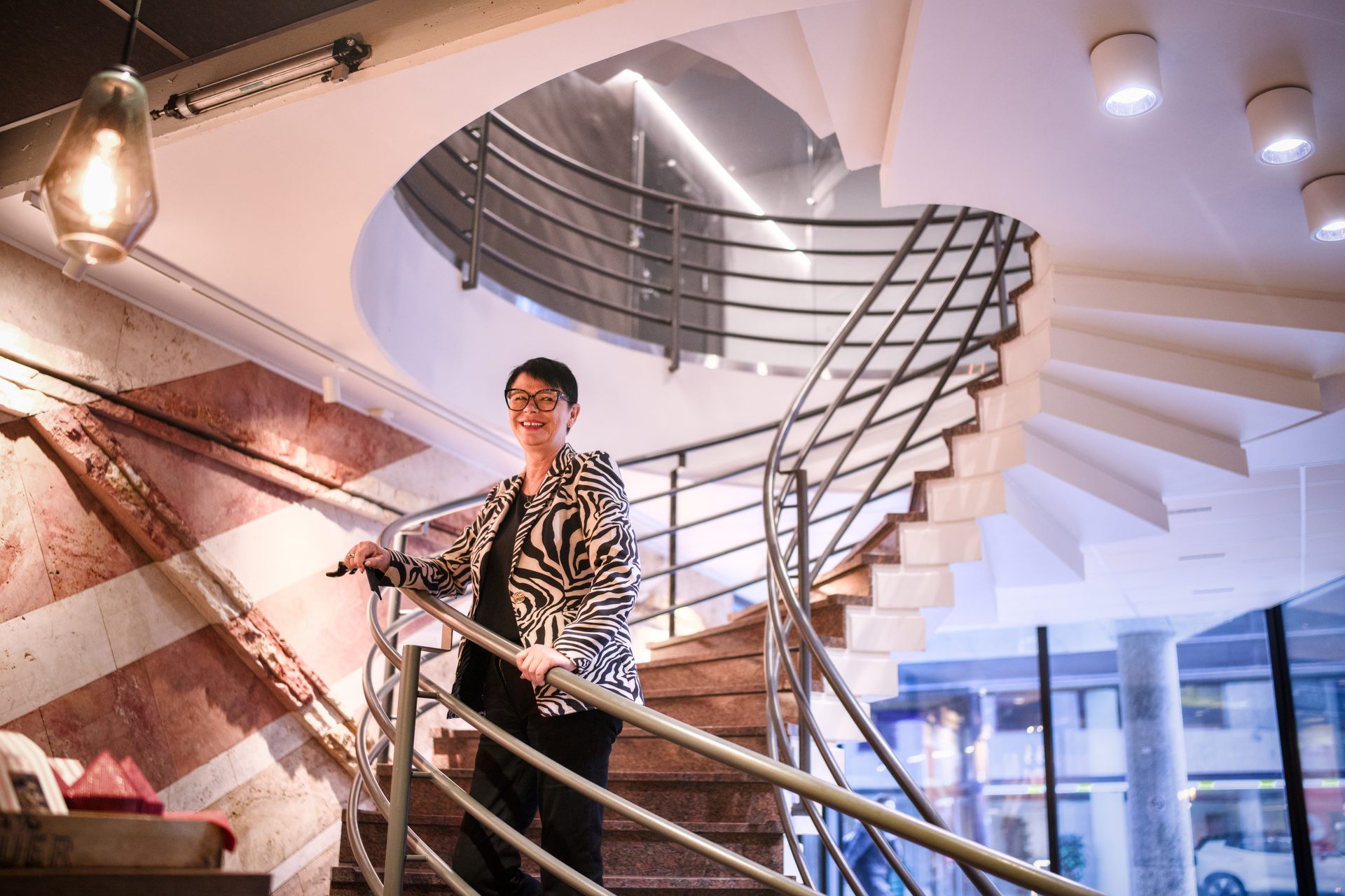 Smiling woman in a zebra jacket stands on a modern spiral staircase.