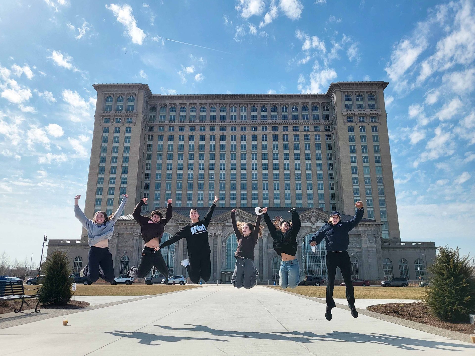 Six people jumping in front of a grand, multi-story building under a blue, cloudy sky.