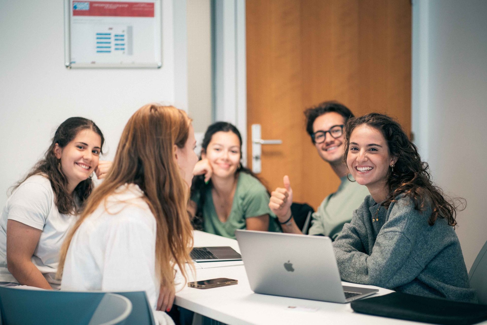 Five smiling students, three girls and two boys, working together at a table with laptops in a bright room.