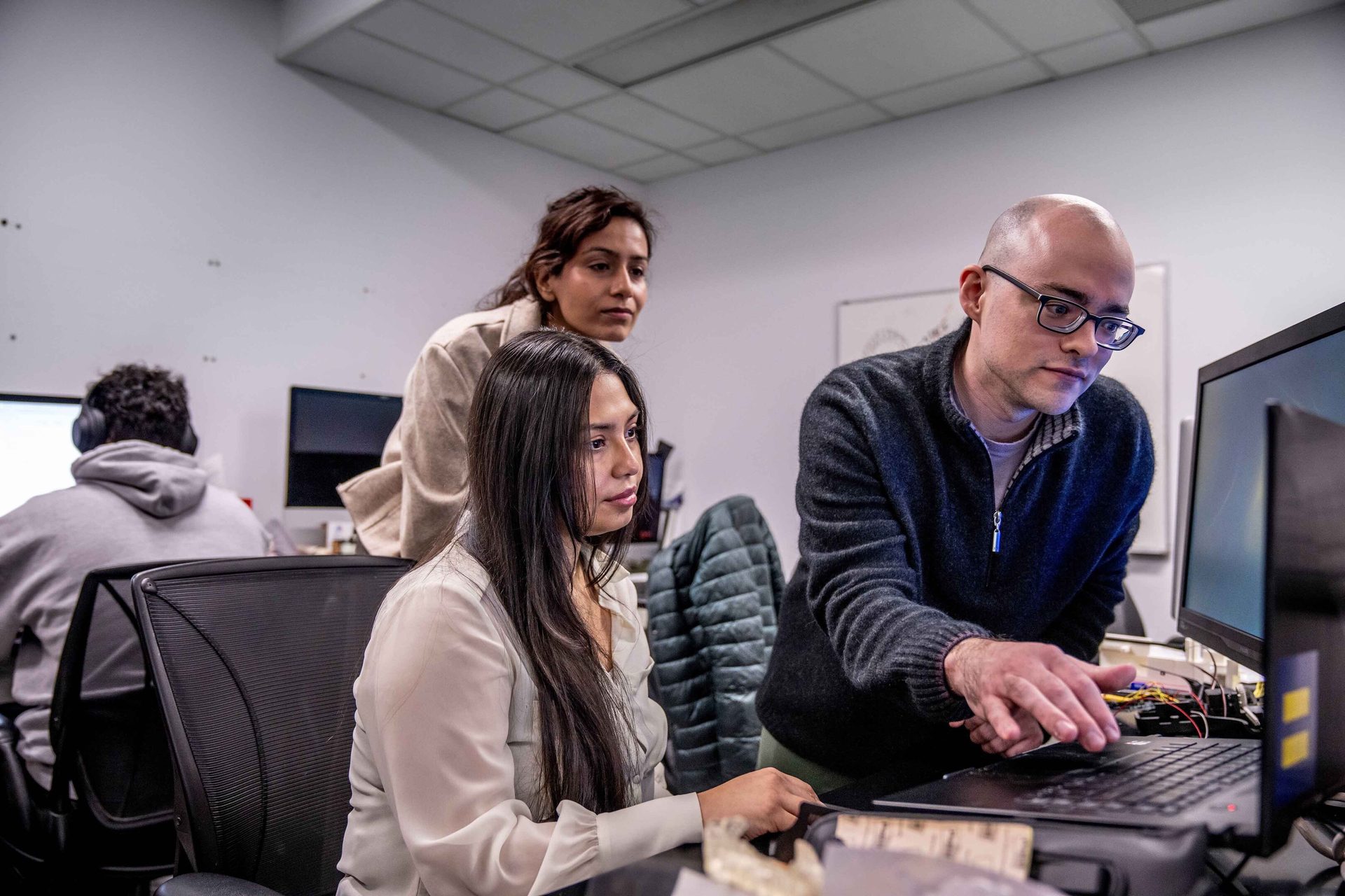 Three colleagues collaborate on a computer, with one man pointing at the screen in an office.