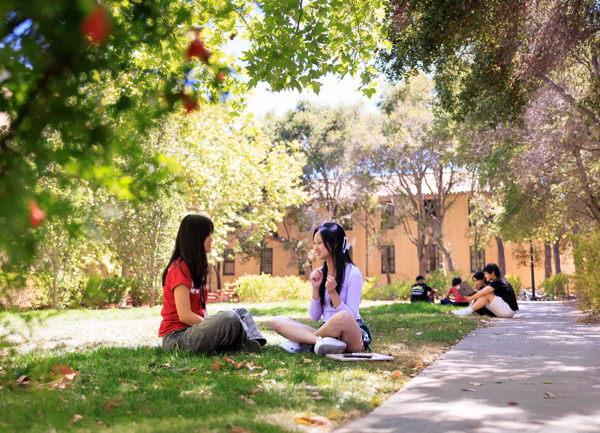 Two women sit on grass, conversing, with trees and a building in the background.