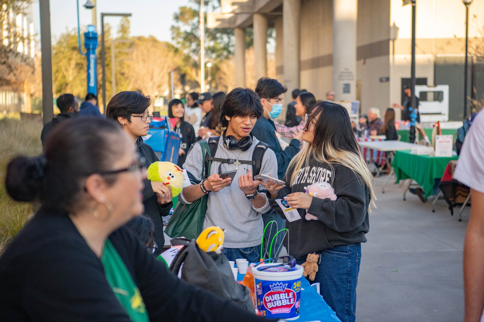 A group of young adults, mostly Asian, interacting at an outdoor event or fair, holding plushies and phones.