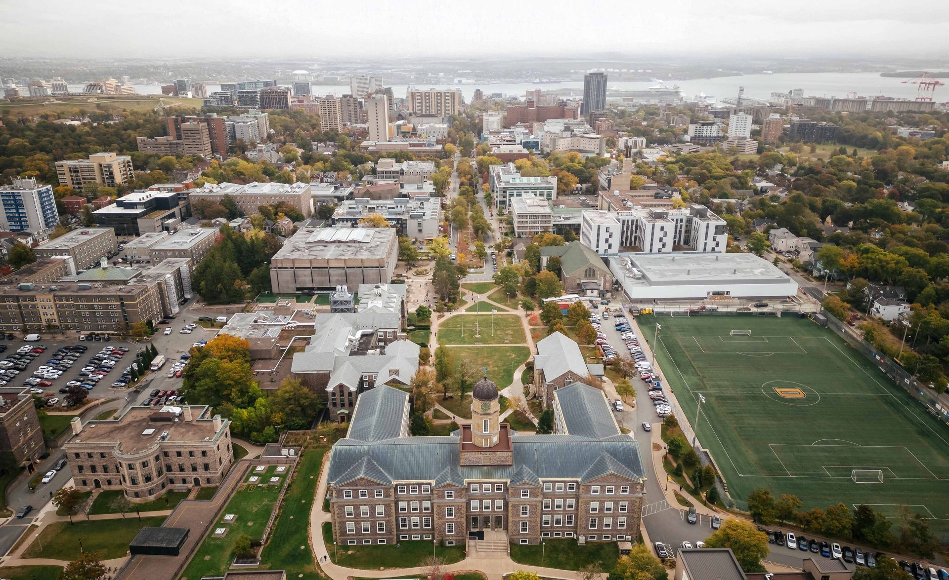Aerial view of a university campus with a large stone building, sports field, and city skyline.