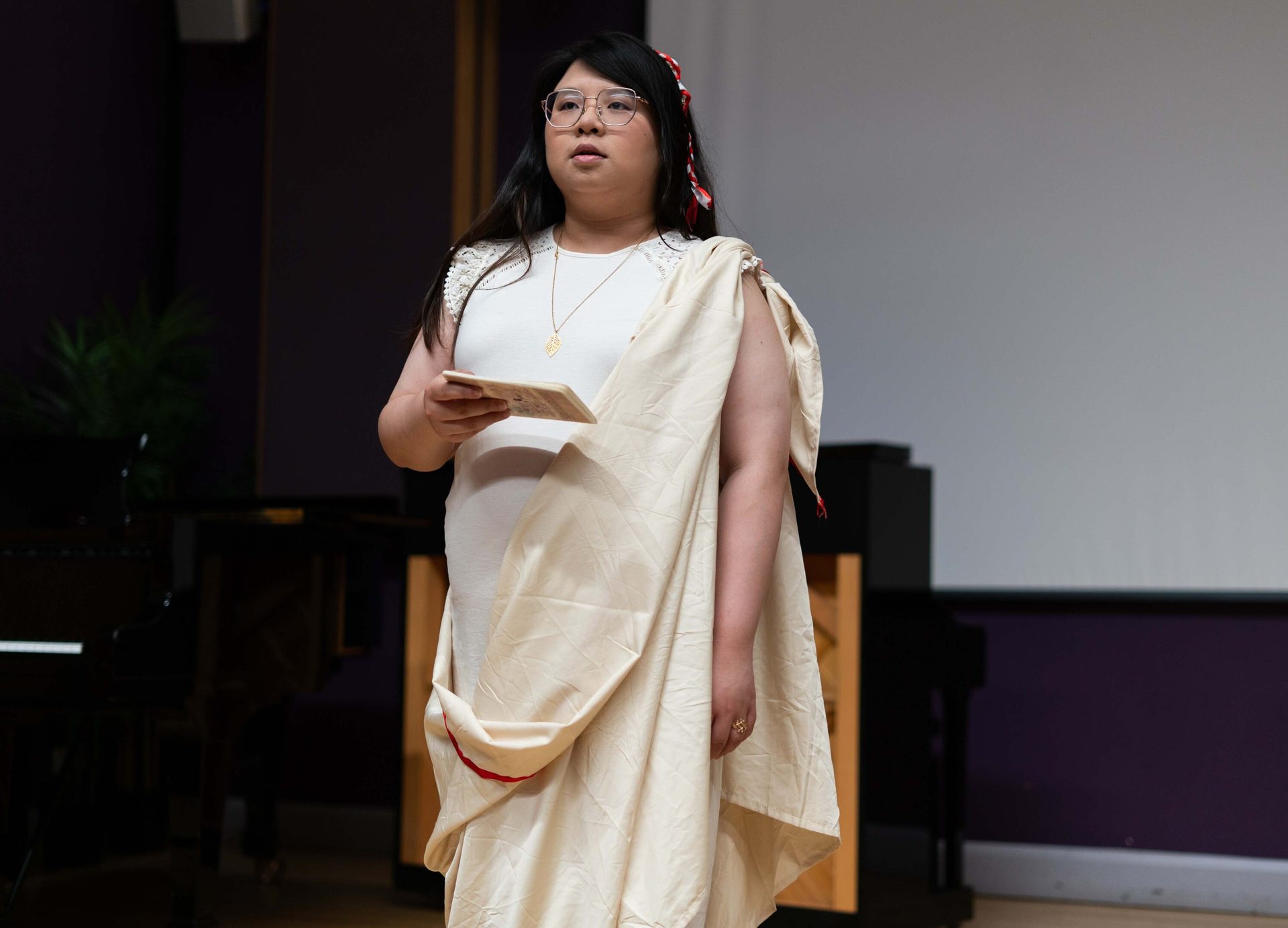 Woman in white dress and draped fabric, glasses, holding small book, standing.