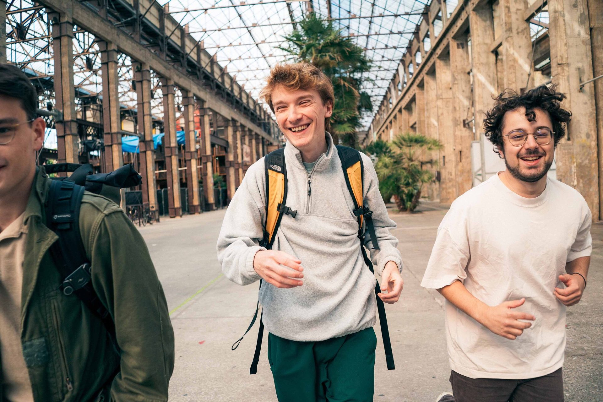 Three young men smiling and walking together in an industrial-style building with a glass roof.