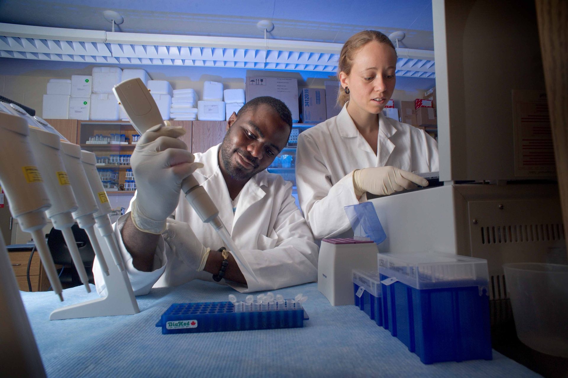 Two scientists in a lab, one pipetting, the other at a machine.