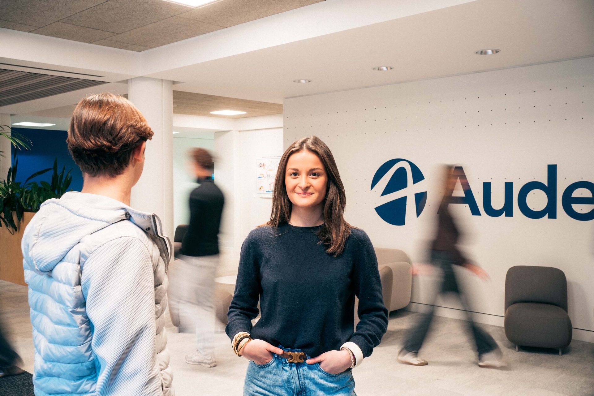 Smiling woman in an office, with the Aude logo on the wall and blurred people in background.