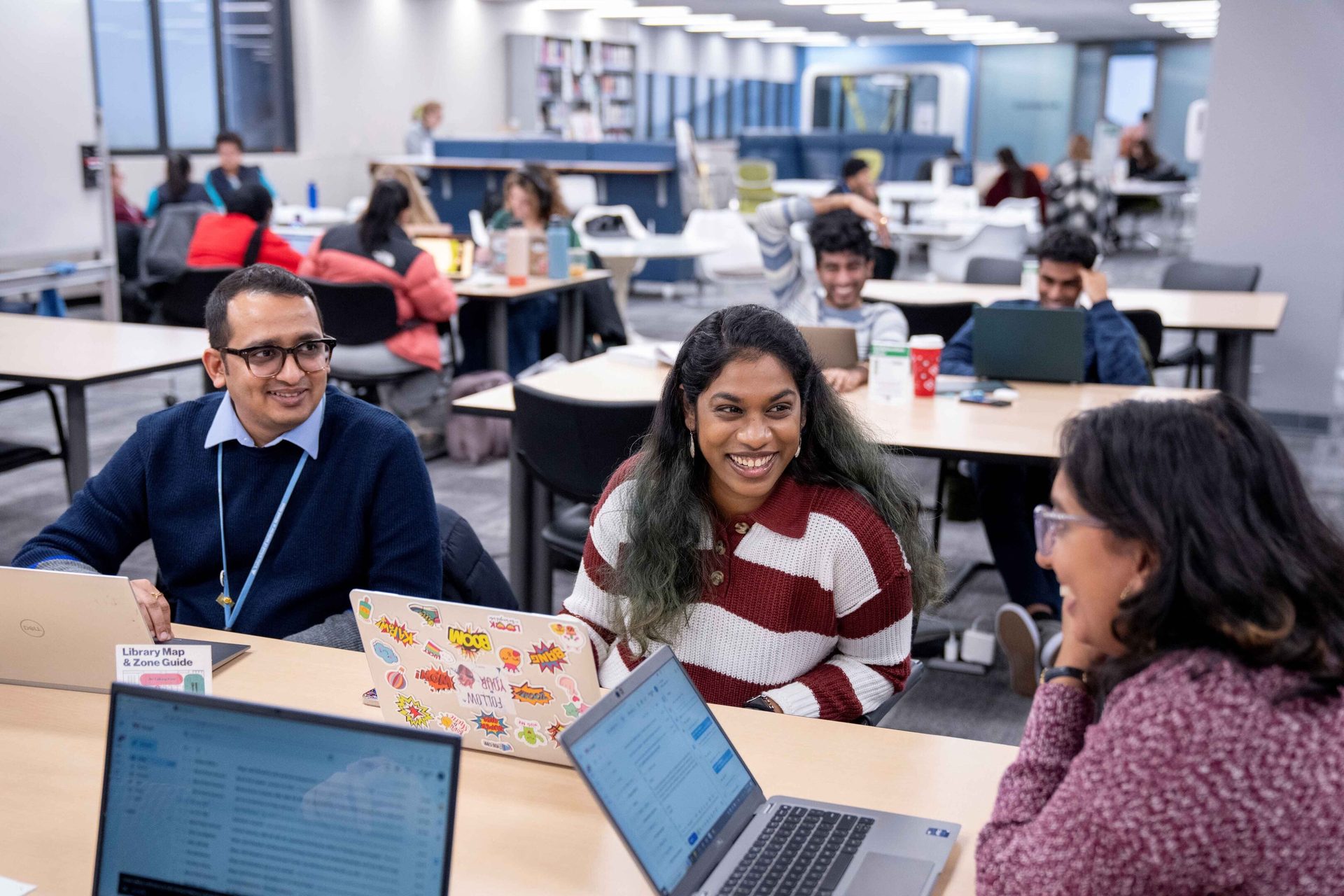 Diverse students smiling, working with laptops in a bright, modern study space.