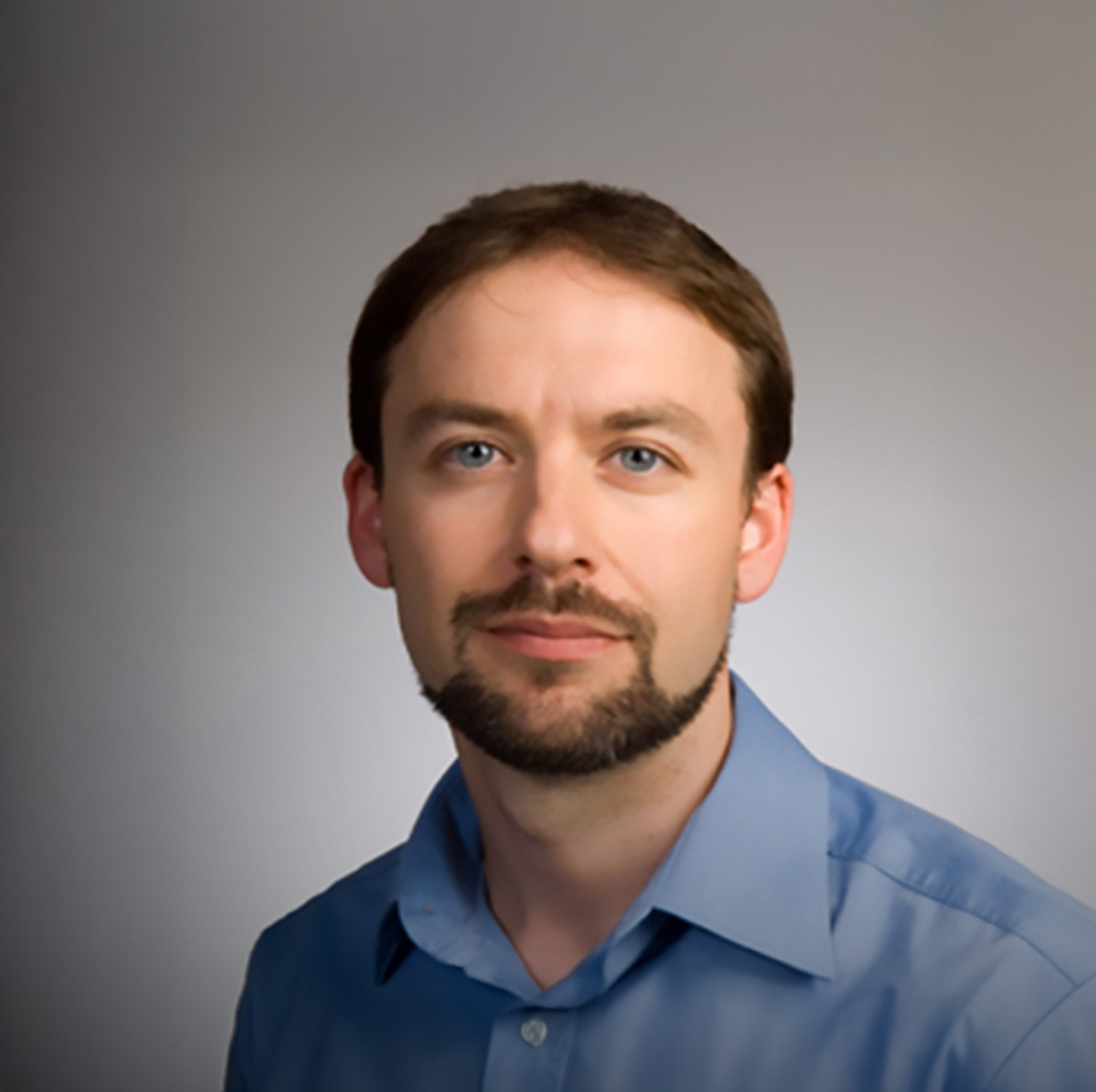 A man with brown hair, blue eyes, and a goatee, wearing a blue shirt against a grey background.