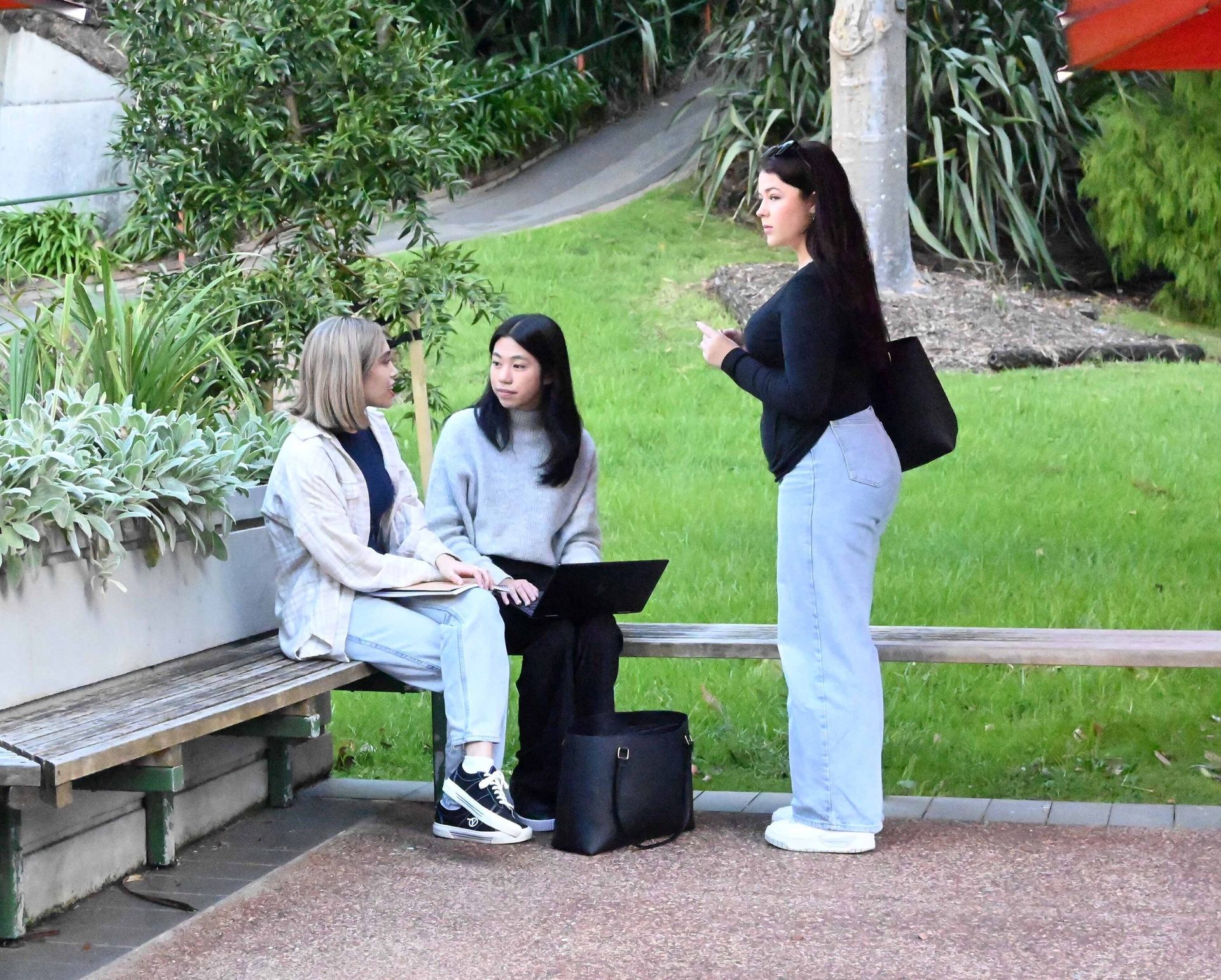 Three women talking outdoors: two on a bench with a laptop, one standing.