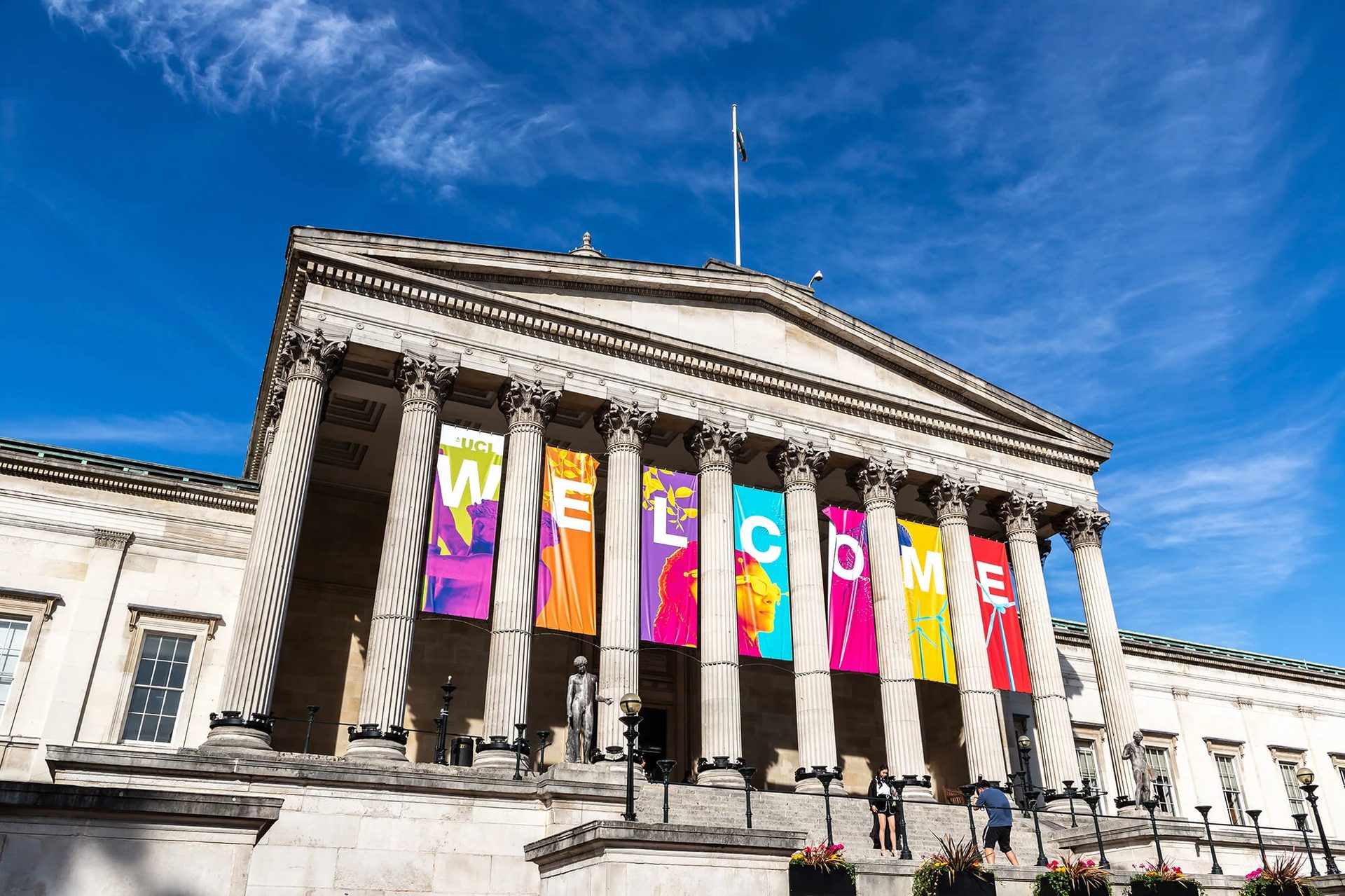 A neoclassical building with columns adorned with colorful 'WELCOME' banners under a blue sky.