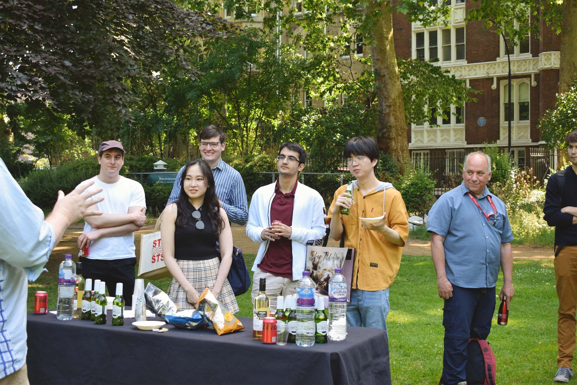 Diverse group of people socializing around a refreshment table outdoors at a sunny gathering.