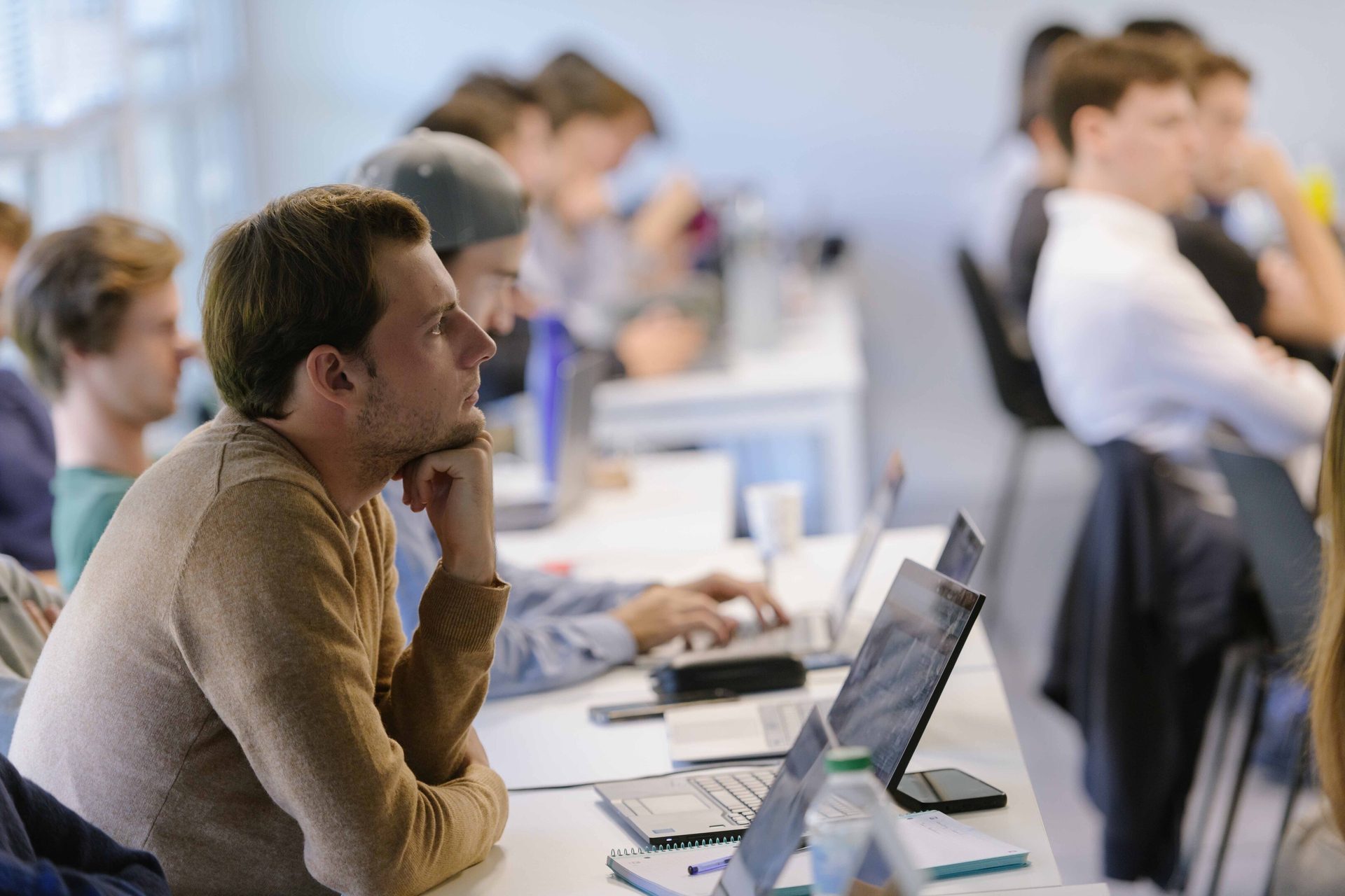 Young man with chin on hand intently watching, surrounded by other students and laptops in a classroom.