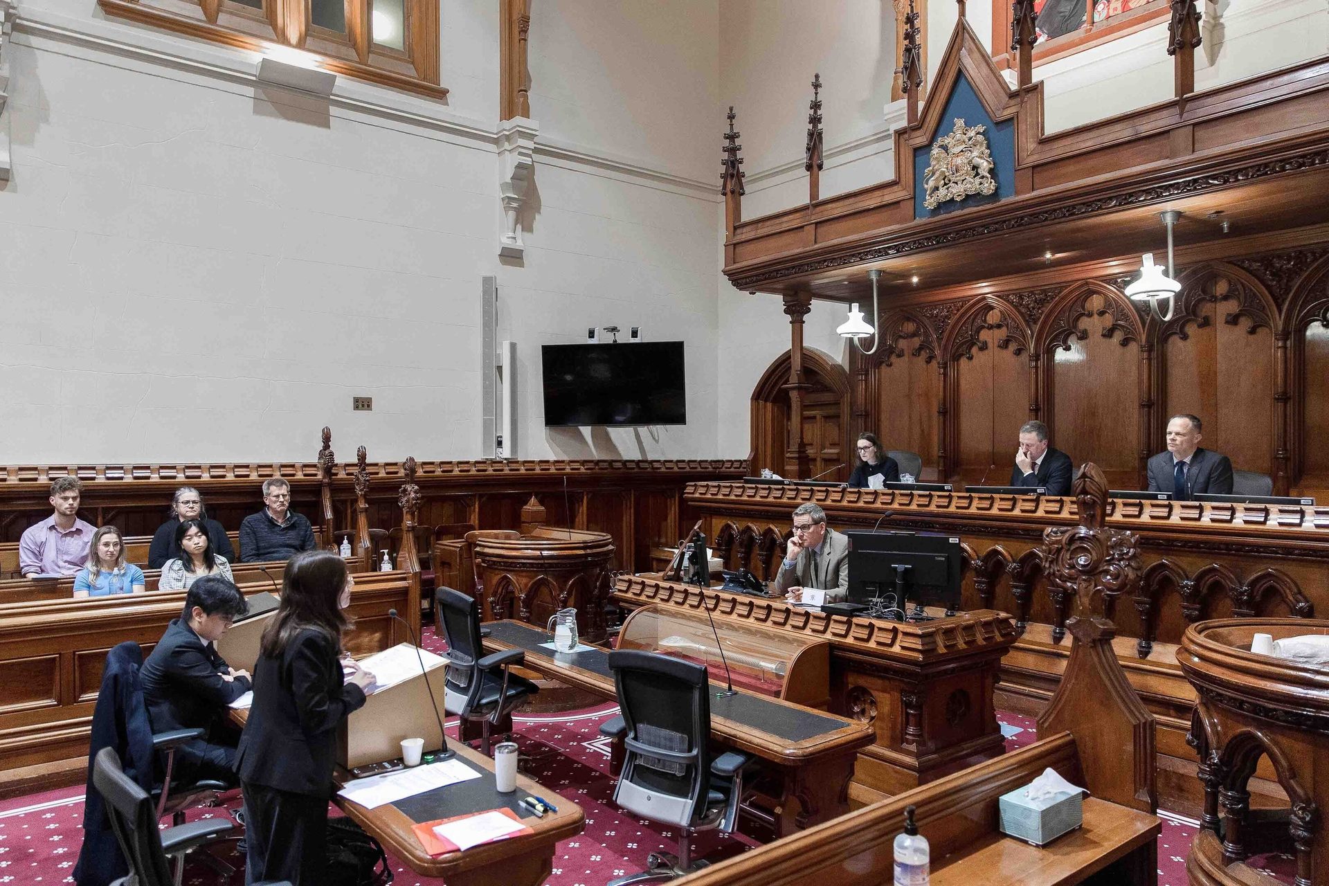 Formal meeting in an ornate wood-paneled chamber with a speaker and attendees.