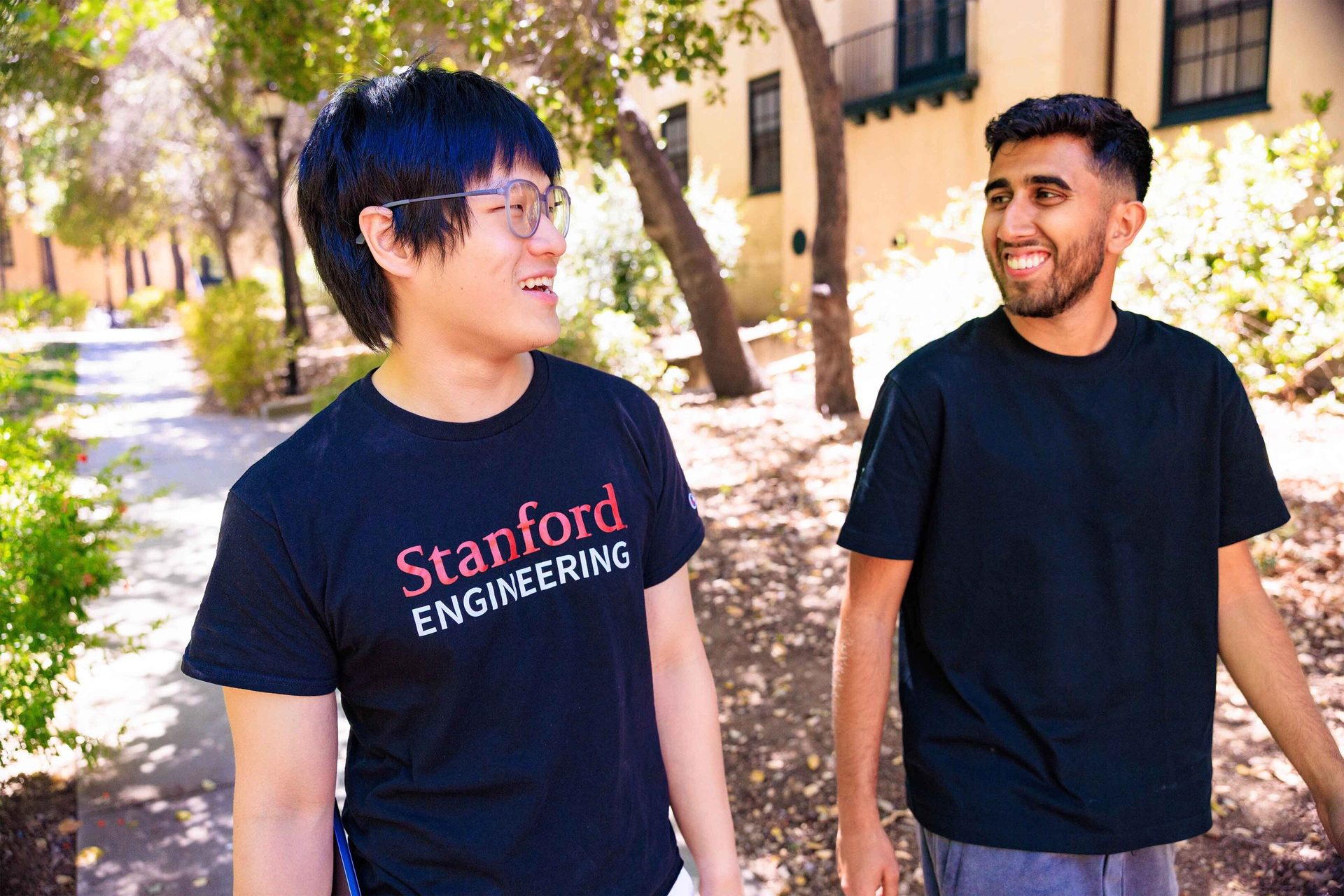 Two smiling male students, one in a Stanford Engineering shirt, walking outdoors on campus.