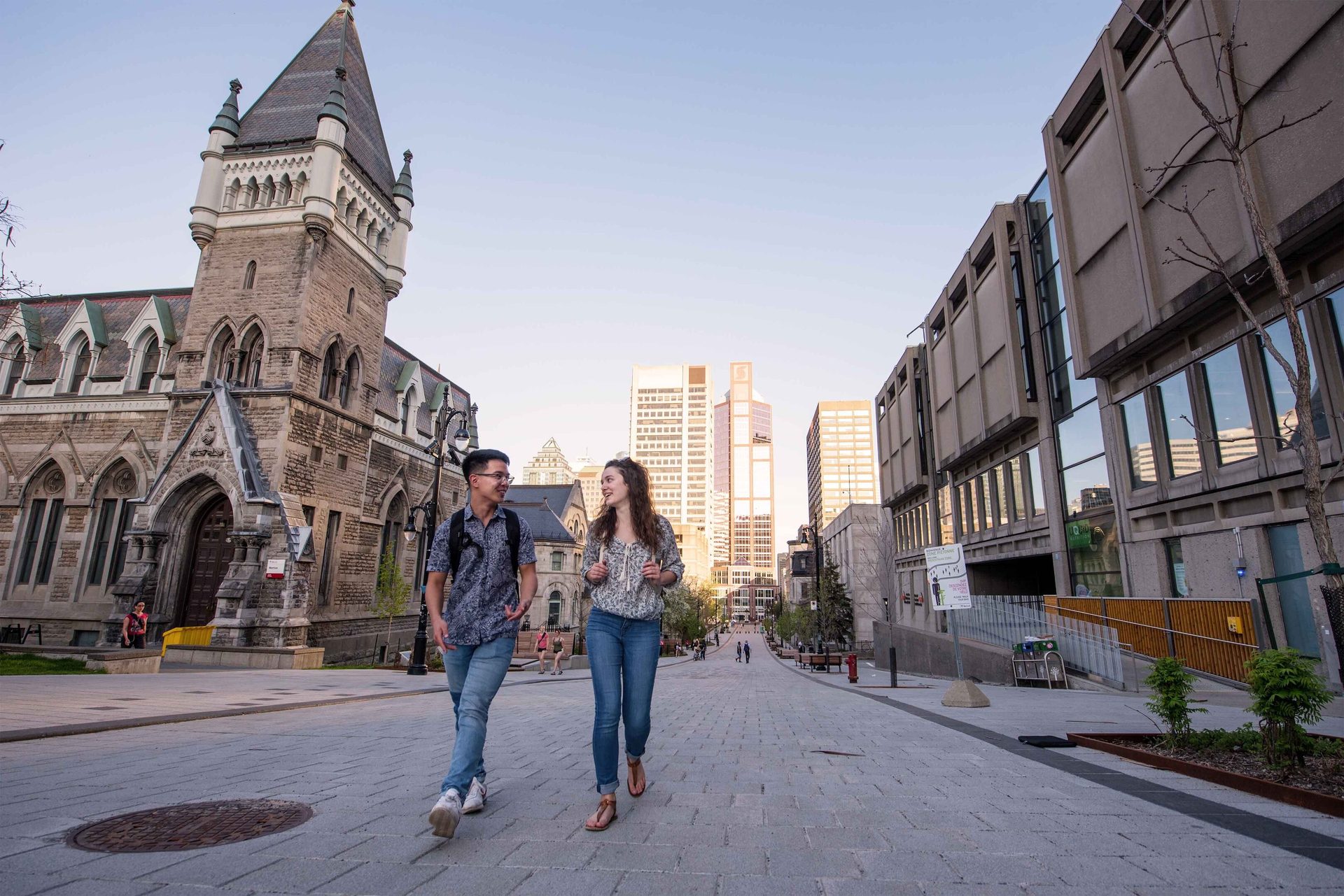 Two young adults walk and talk on a stone-paved street, flanked by historic and modern buildings.