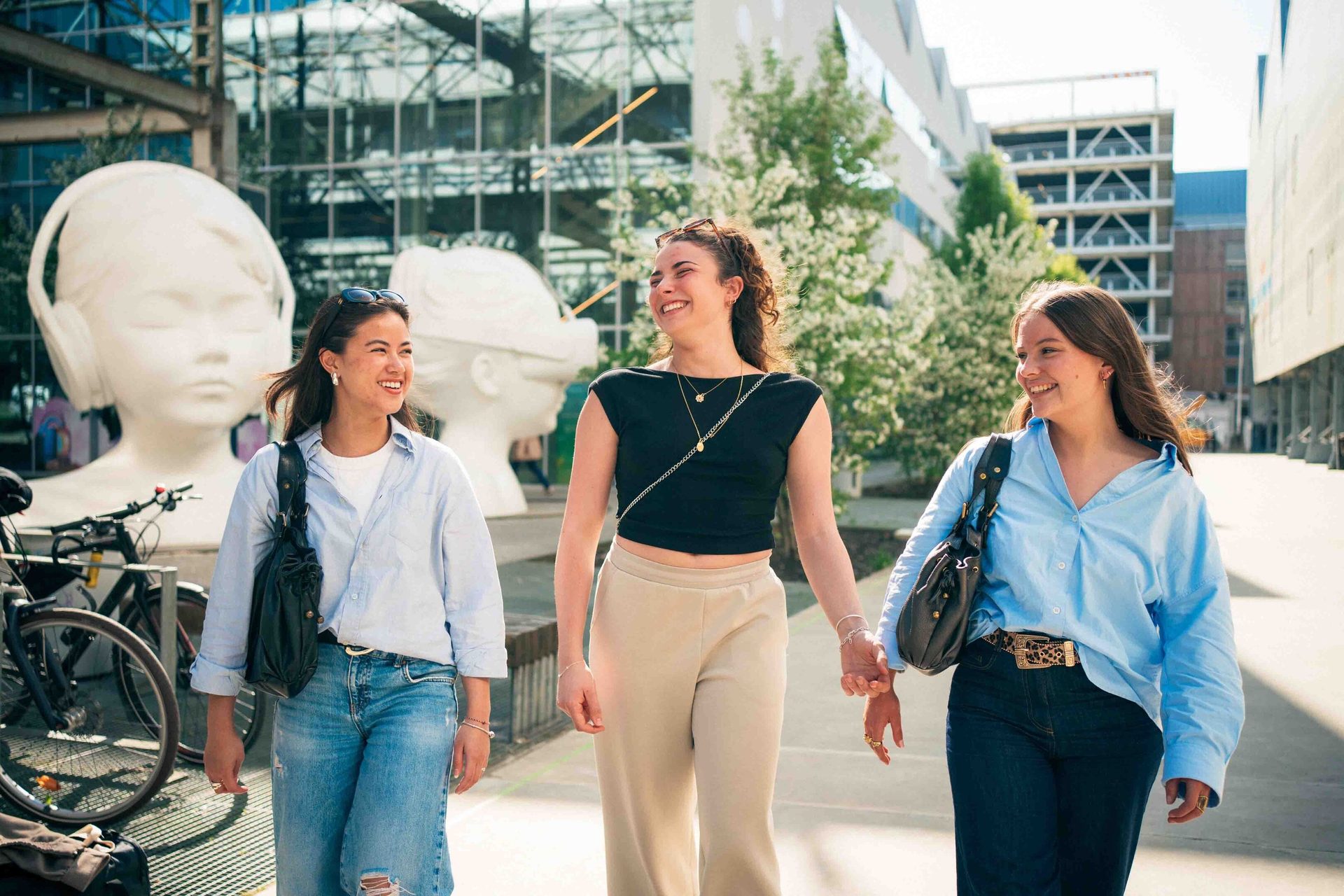 Three smiling young women walk hand-in-hand outdoors in a city, with large white sculptures in the background.