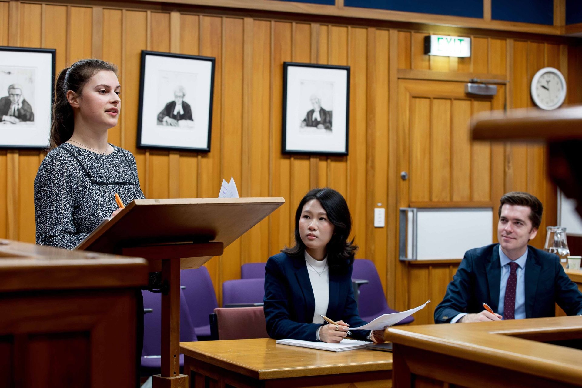 Young woman speaking at a podium in a courtroom setting, two people listen at tables.