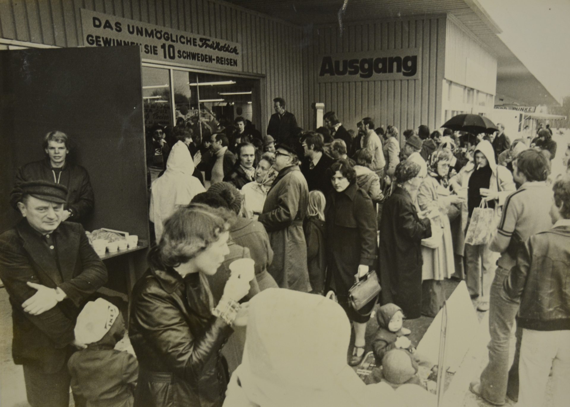 A large crowd gathers outside a building with an "Ausgang" (Exit) sign, possibly for an event or sale.