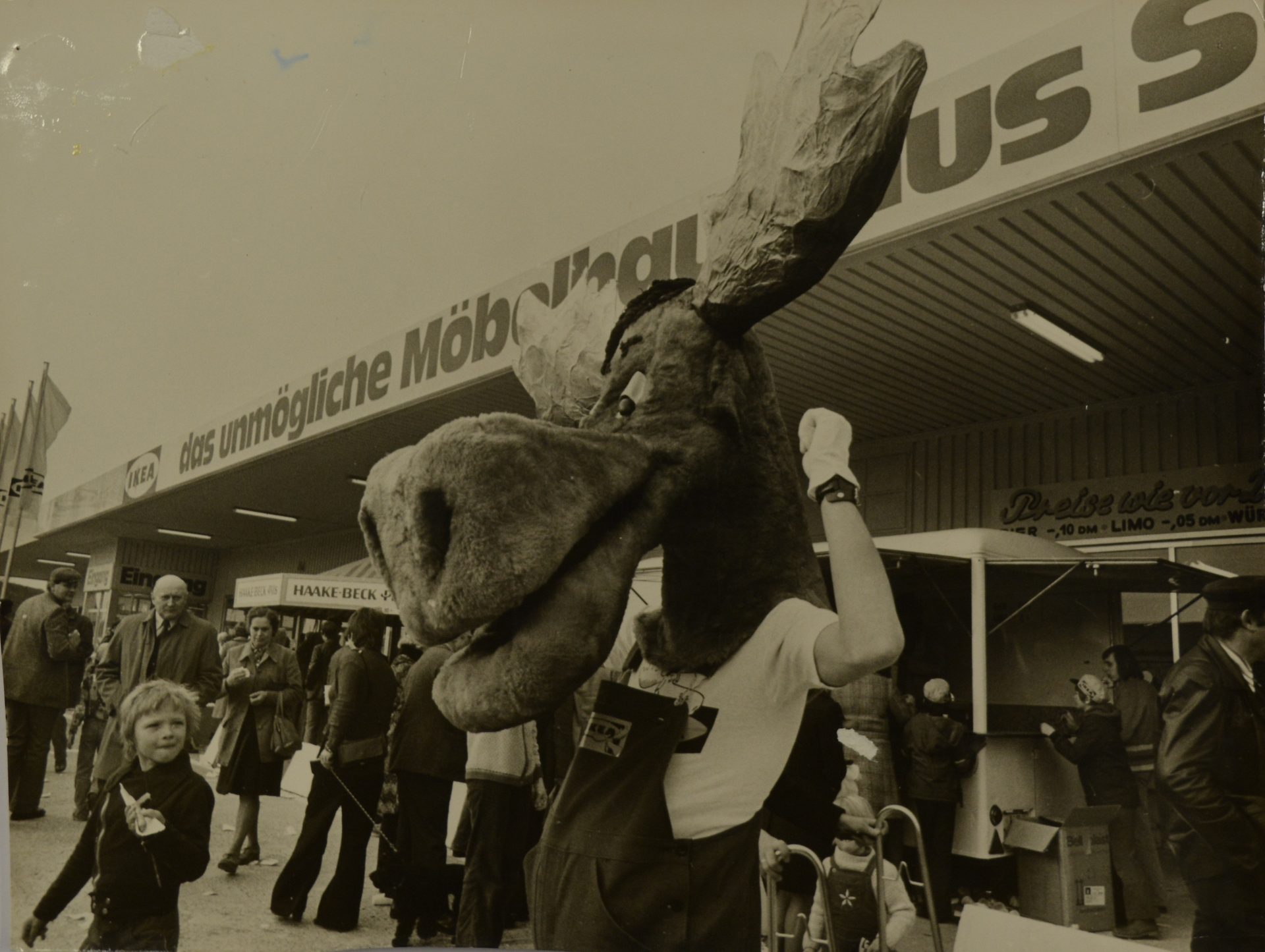 A person in a large moose mascot costume stands near an IKEA store entrance, with a child in the foreground.