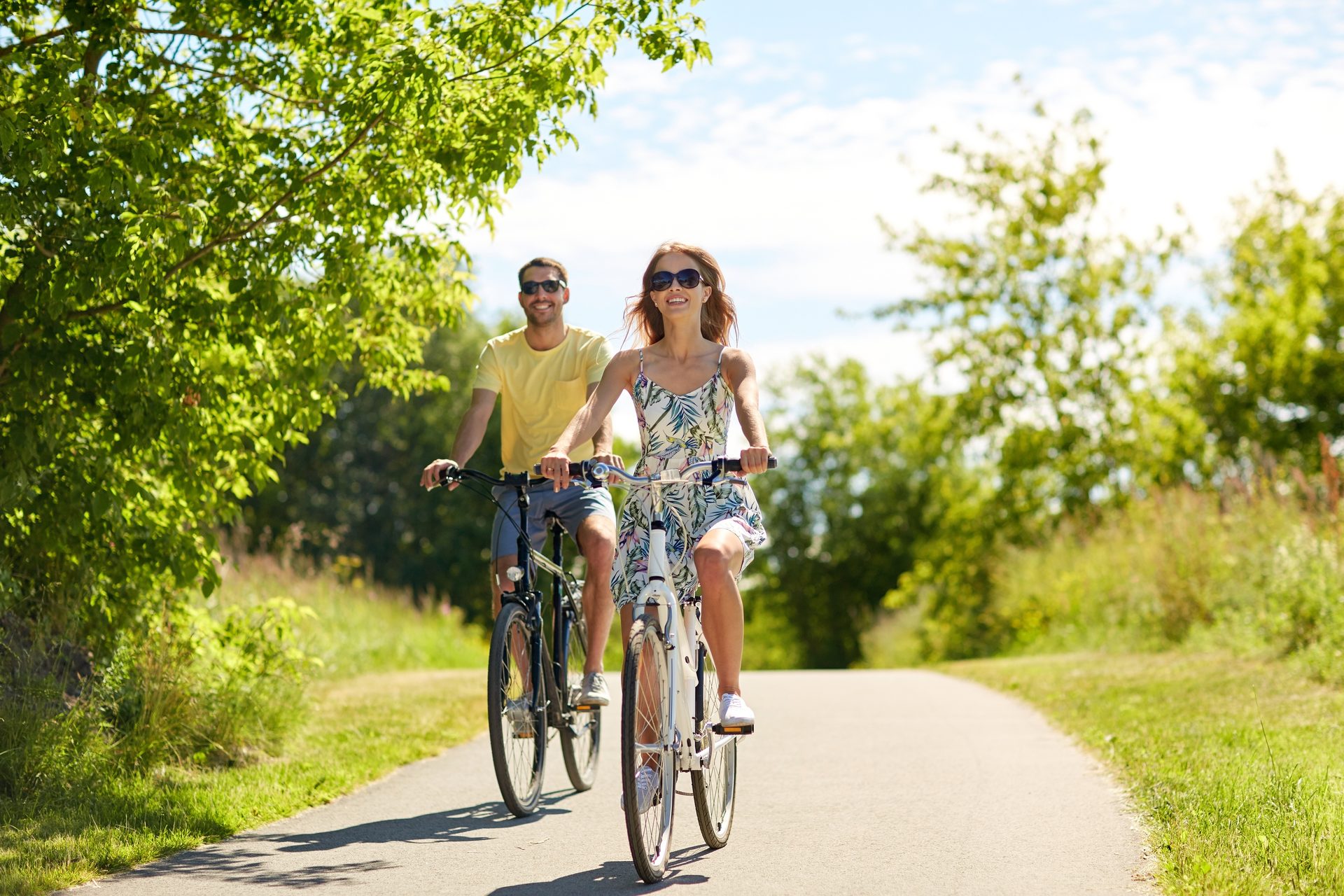 A happy couple on bicycles rides along a sunny paved path surrounded by green trees, enjoying their time.